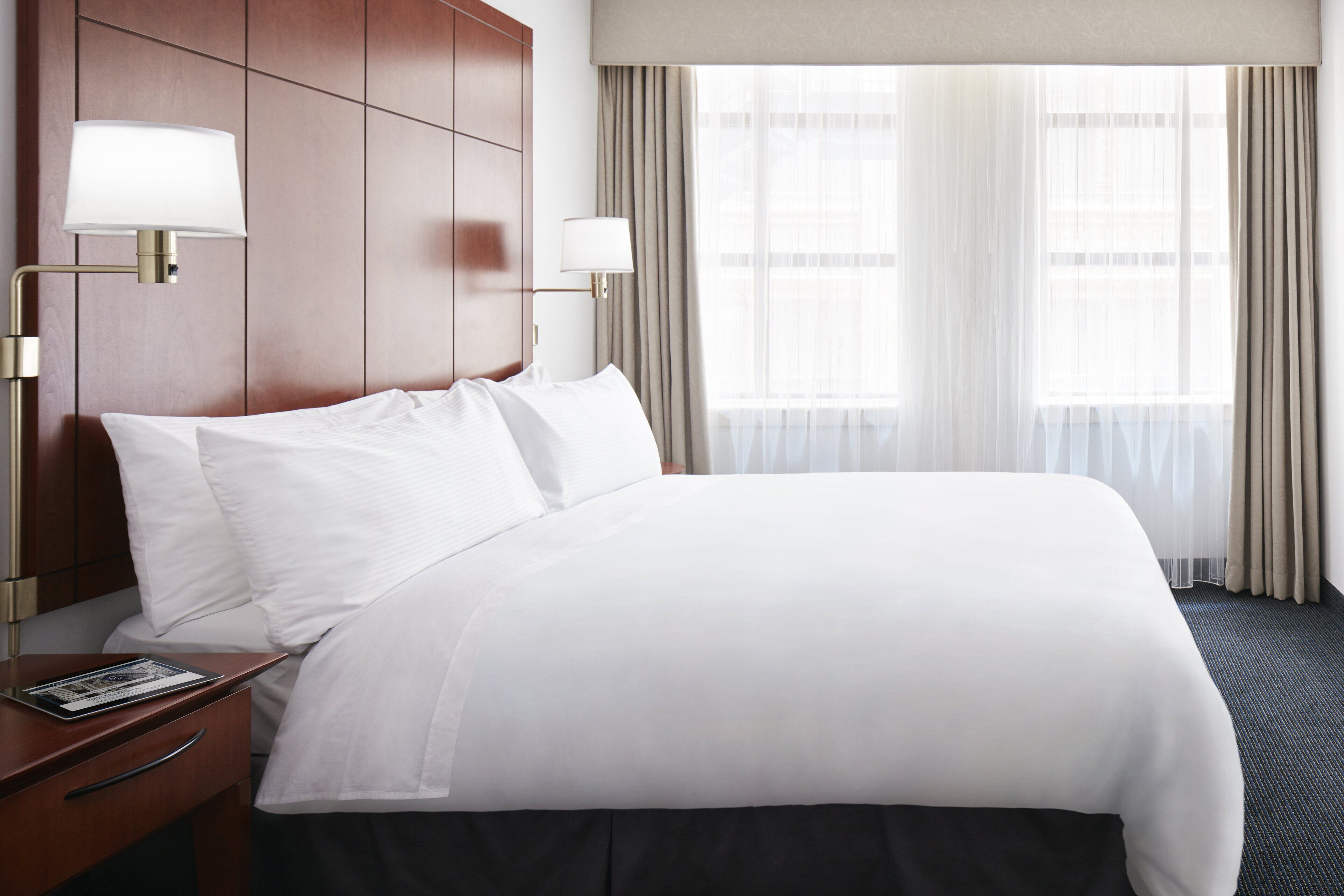 Una habitación de hotel con una cama bien hecha, un cabecero de madera, dos lámparas de noche, una mesita de noche y una ventana con cortinas.