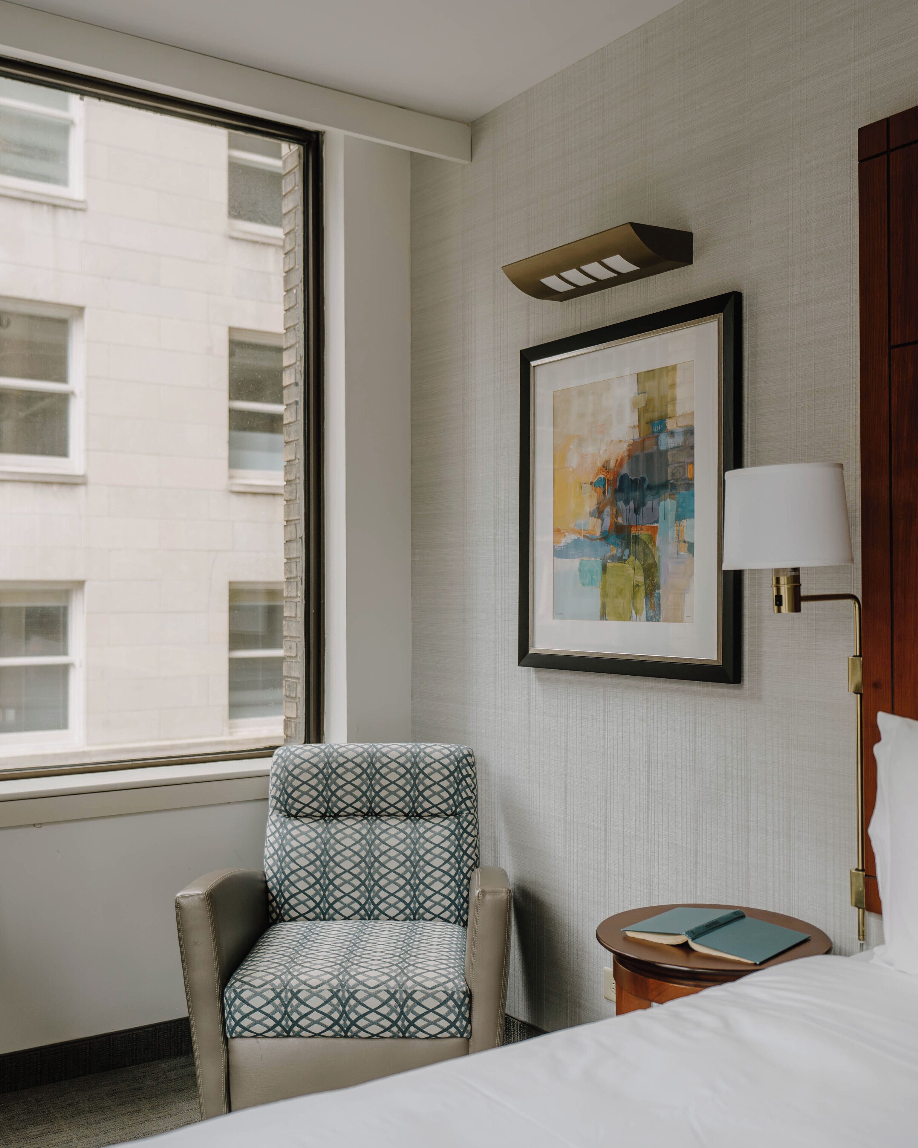 Quarto de hotel com uma janela grande, uma pintura abstrata emoldurada, uma poltrona estampada, uma mesa de cabeceira com um livro e um abajur.