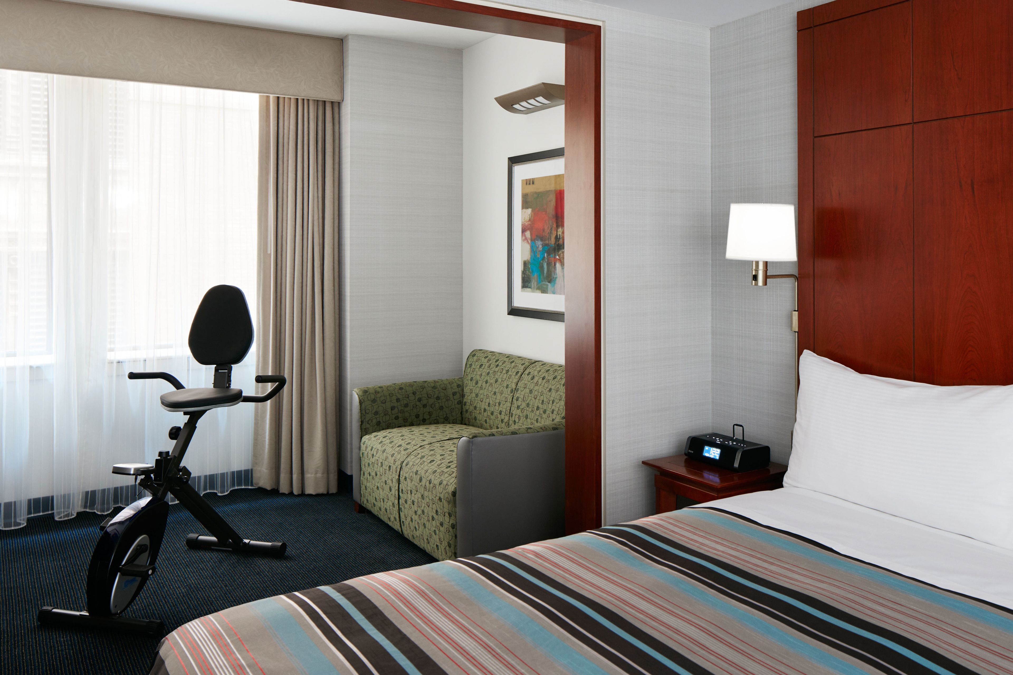 A hotel room with a bed, a desk with a lamp, a chair, an exercise bike, and a painting on the wall.