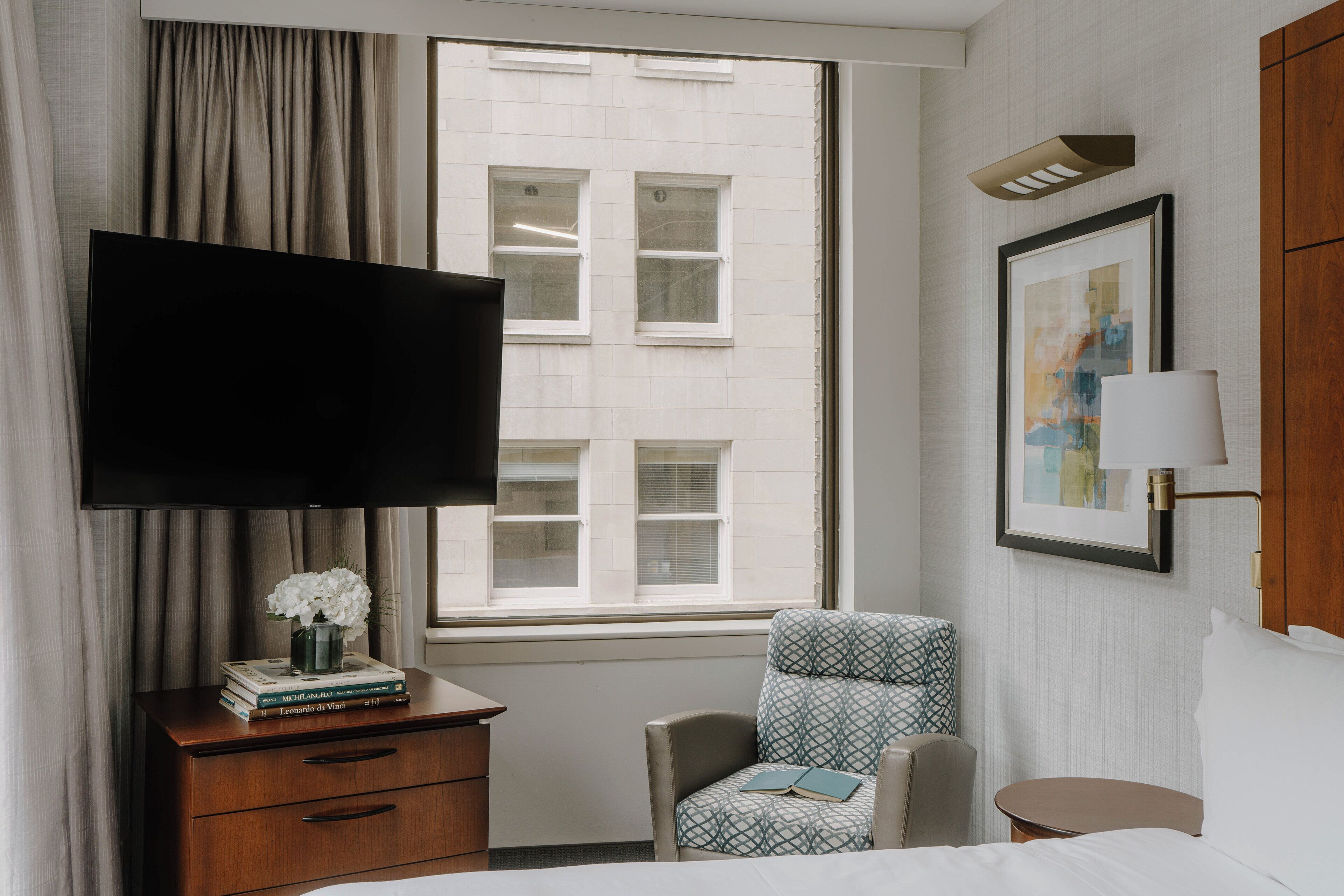 A hotel room with a bed, a desk with a flat-screen TV, a chair, a small table, and a window with a view of buildings.