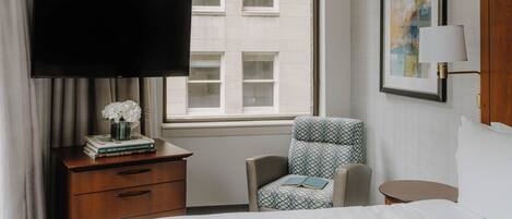 A hotel room with a bed, a desk with a flat-screen TV, a chair, a small table, and a window with a view of buildings.