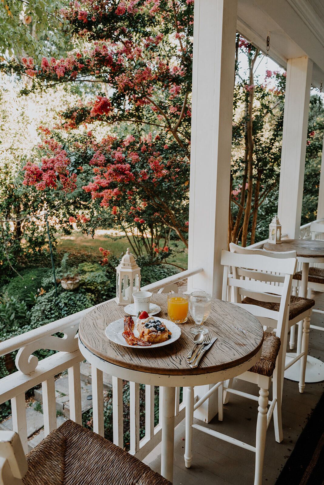 Uma mesa de madeira redonda com um prato de comida, um copo de suco de laranja e uma vela em um alpendre branco com cadeiras brancas.