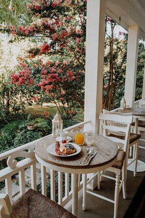 A round wooden table with a plate of food, a glass of orange juice, and a candle on a white porch with white chairs. - Mayhurst Inn (Orange)