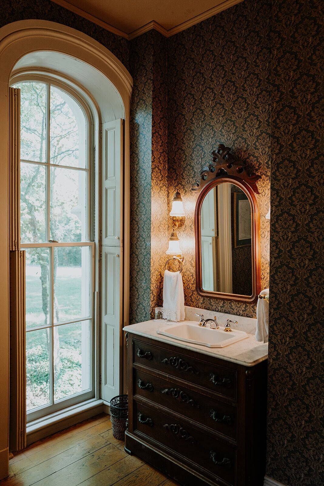 A bathroom with a large arched window, a vanity with a mirror, and a wall with a floral pattern.