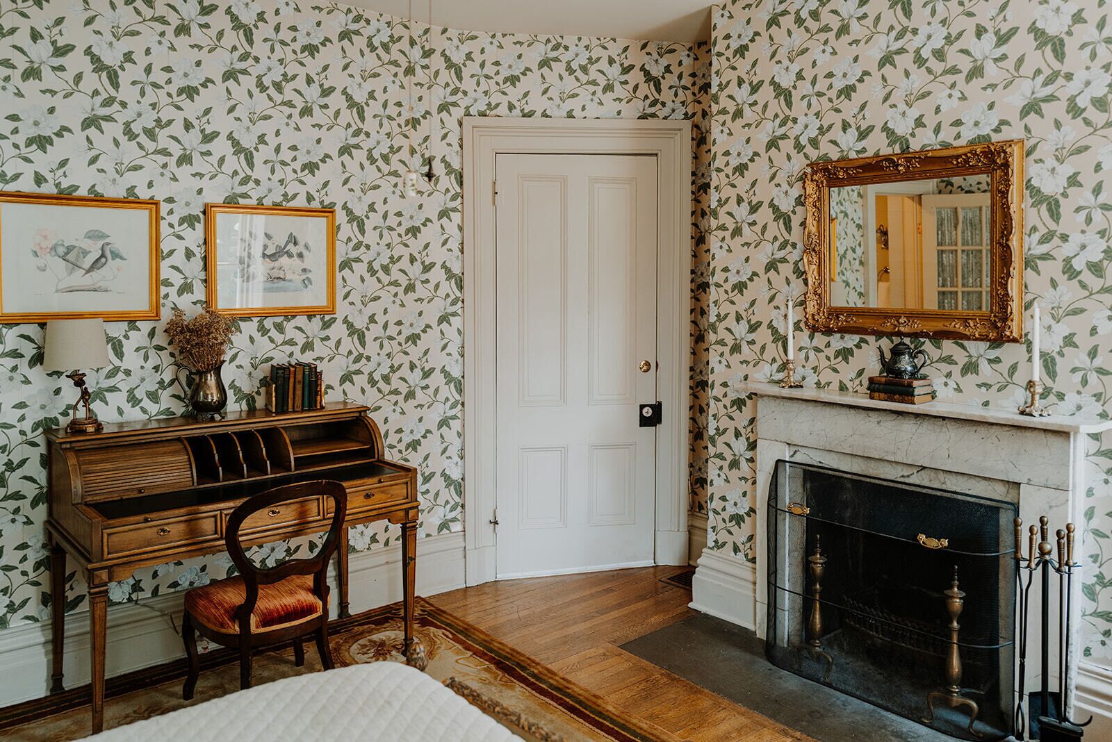 A room with floral wallpaper, a white door, a fireplace with a mantel, and a wooden desk with a chair.