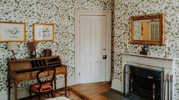 A room with floral wallpaper, a white door, a fireplace with a mantel, and a wooden desk with a chair.