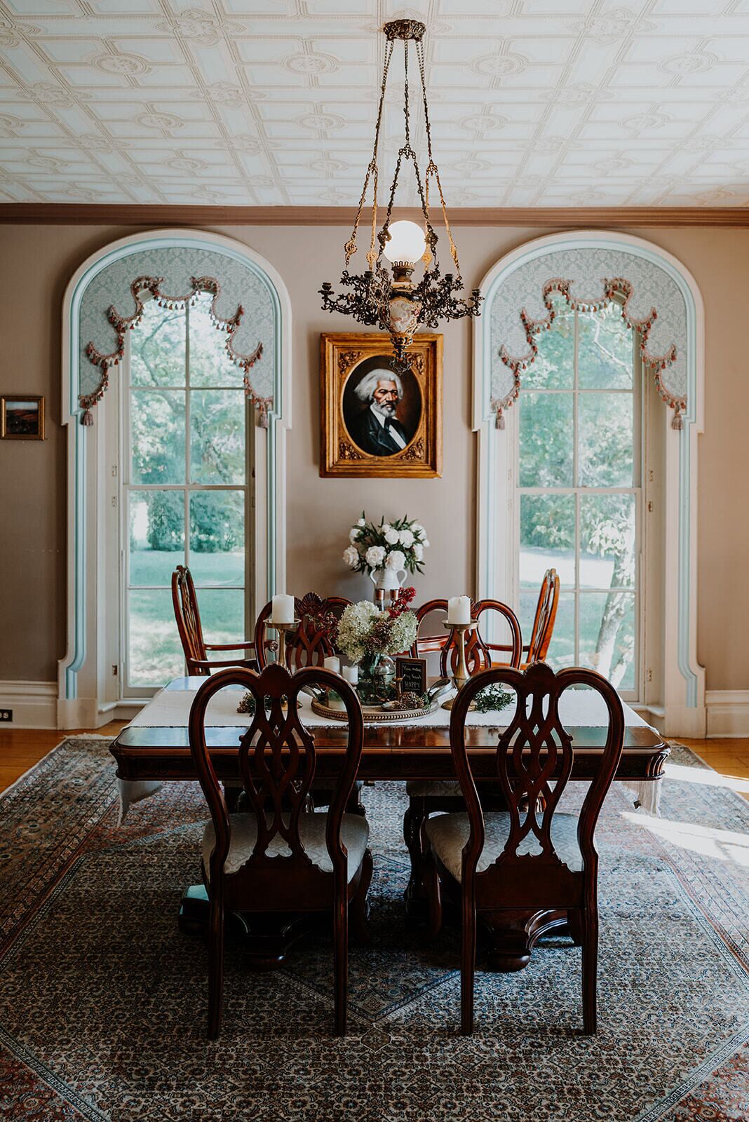 A dining room with a table set for six, surrounded by ornate chairs, a chandelier, and a framed portrait on the wall.