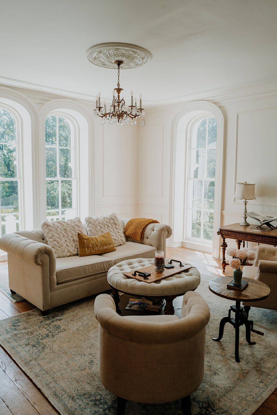 A room with a sofa, ottoman, and armchairs, a chandelier, and large windows with a view.
