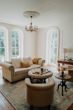 A room with a sofa, ottoman, and armchairs, a chandelier, and large windows with a view. - Mayhurst Inn (Orange)
