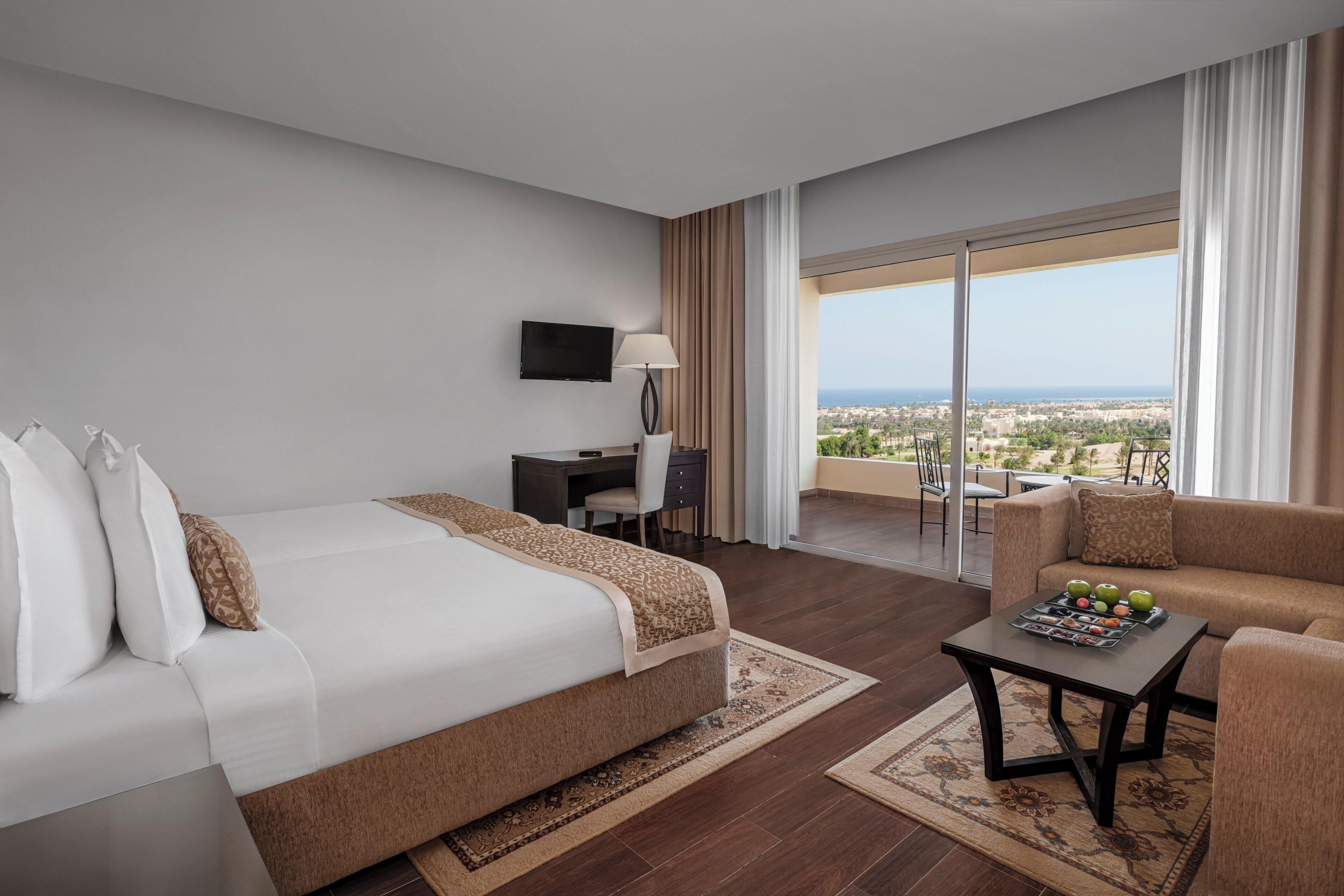 Habitación de hotel con cama, escritorio, sofá, televisor y balcón con vista a la ciudad.
