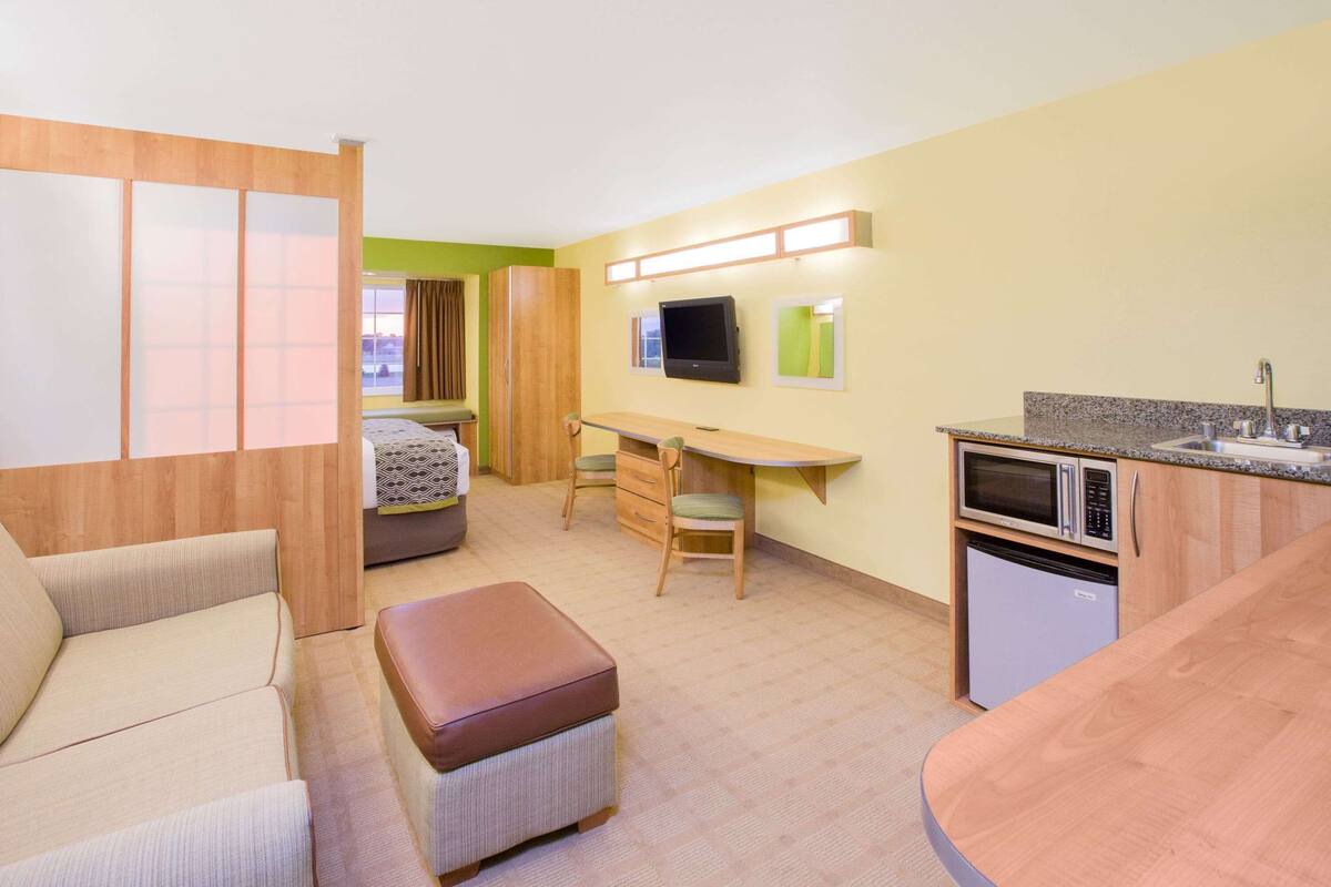 A compact hotel room with a bed, a desk, a kitchenette, and a seating area.