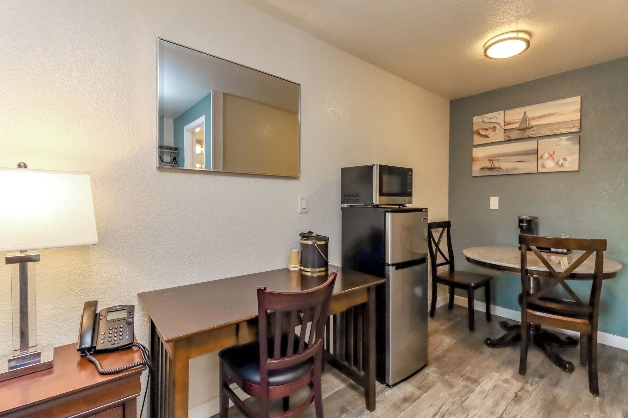 Cocina compacta con refrigerador, microondas y una pequeña mesa de comedor con sillas. Un teléfono está sobre una mesita auxiliar y un espejo colgado en la pared.