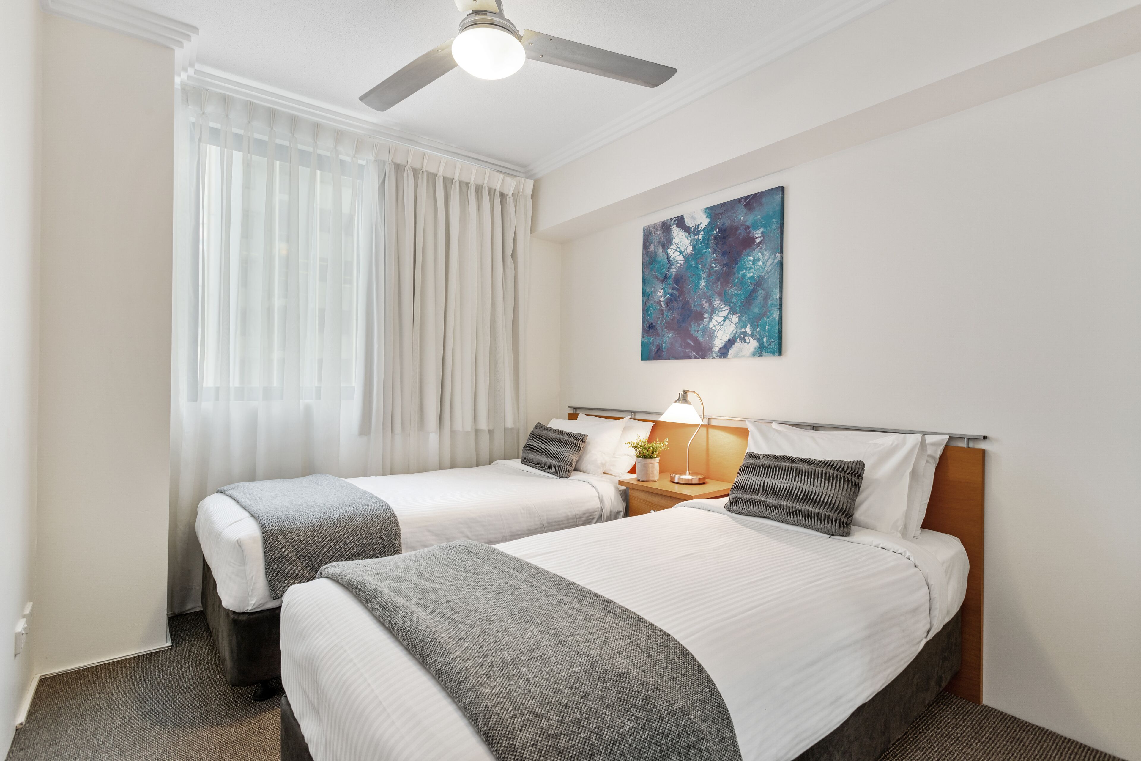 A hotel room with two beds, a ceiling fan, a painting on the wall, and a window with curtains.