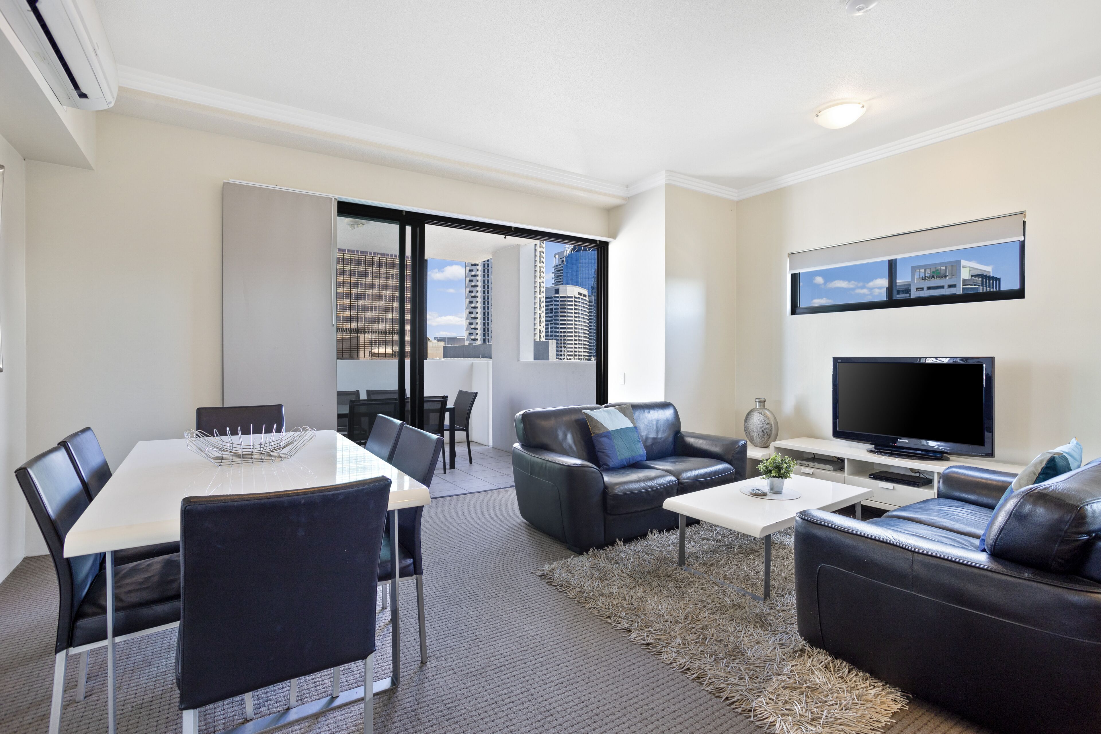 A modern living room with a dining area, a flat-screen TV, and a large window with a city view.