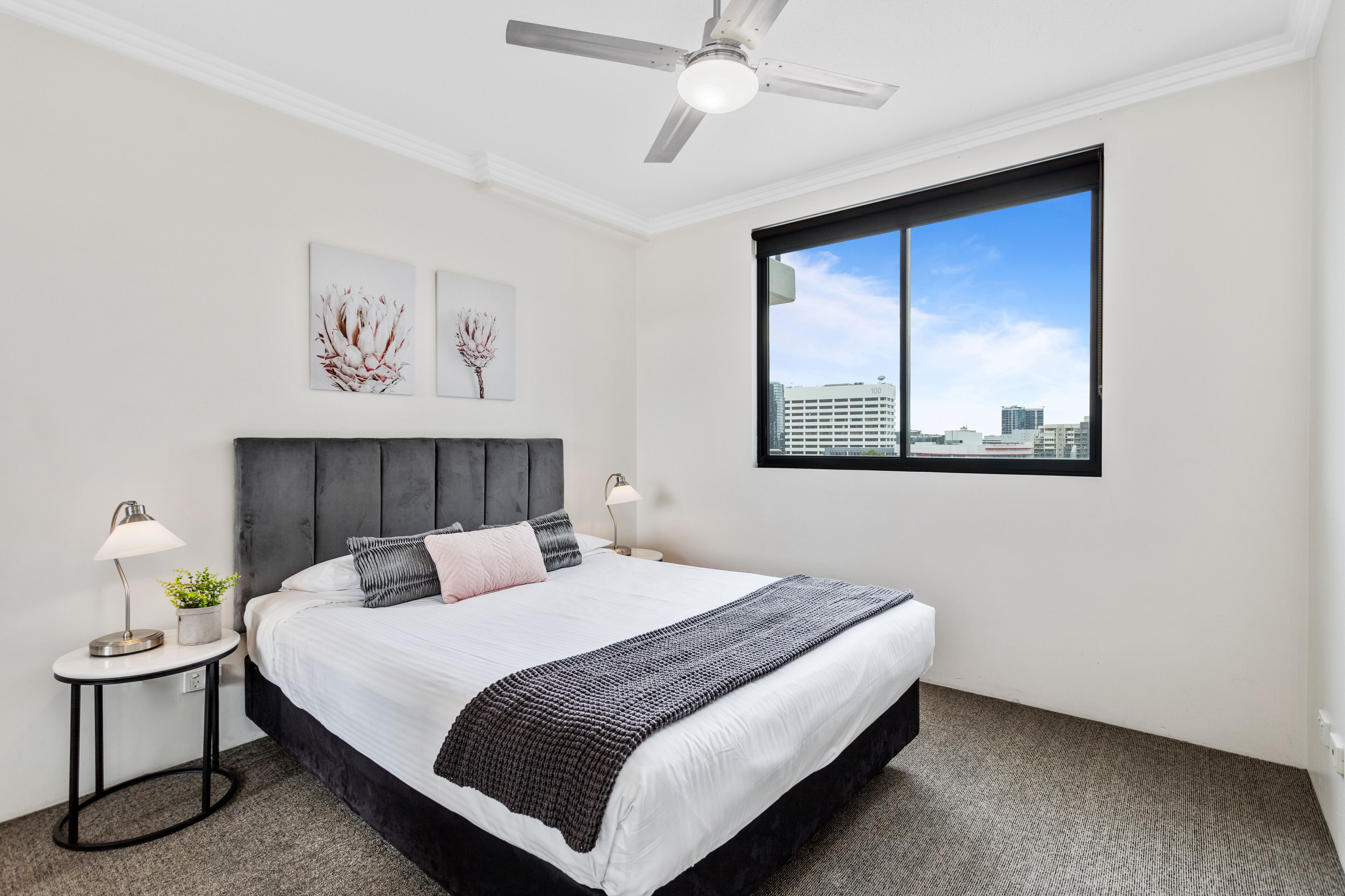 A hotel room with a large bed, a ceiling fan, a window with a city view, and two wall-mounted artworks.