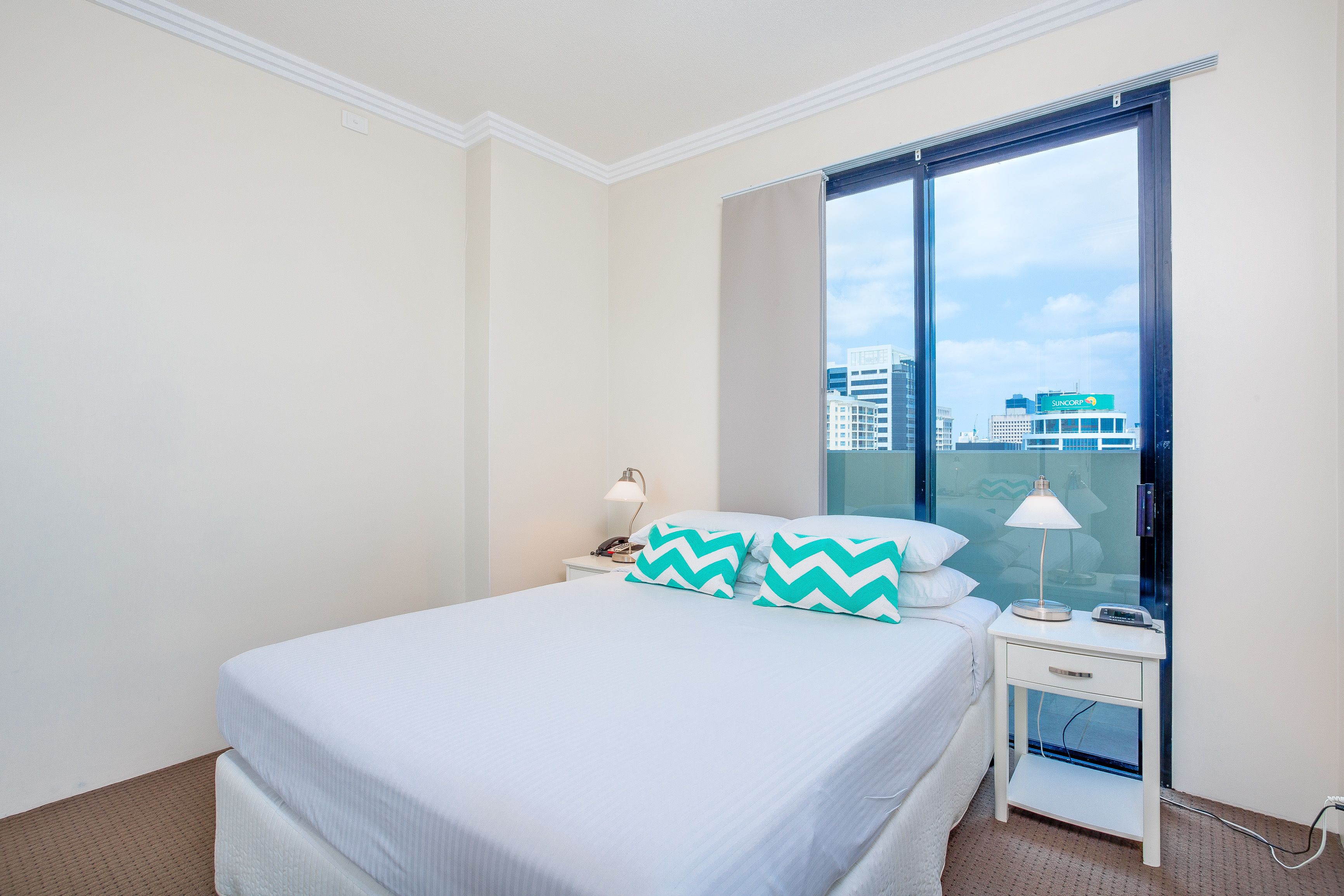 A hotel room with a bed, a nightstand, a lamp, and a window with a city view.
