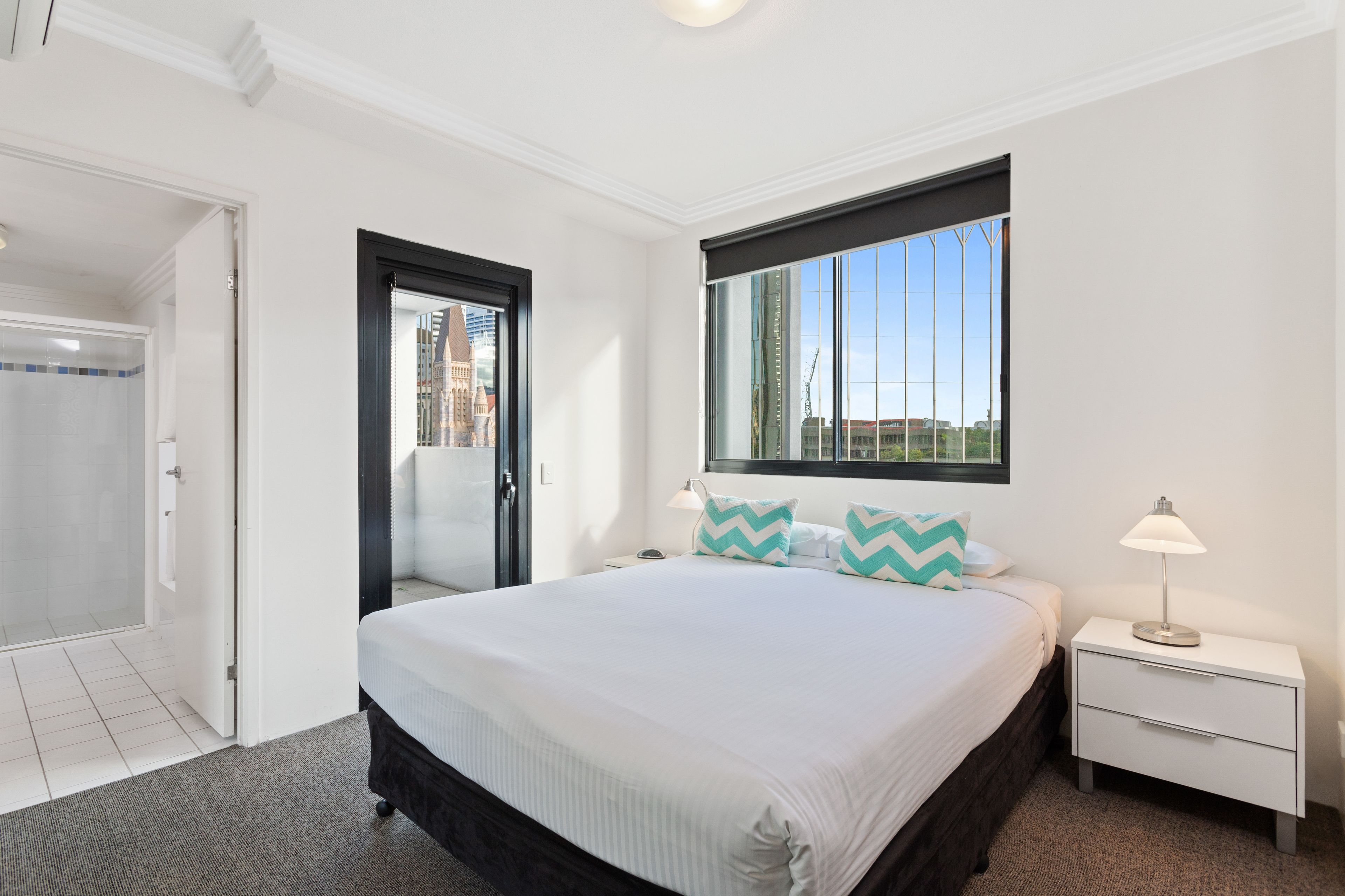 A hotel room with a bed, a nightstand, a window with a view of buildings, and a door leading to a shower area.
