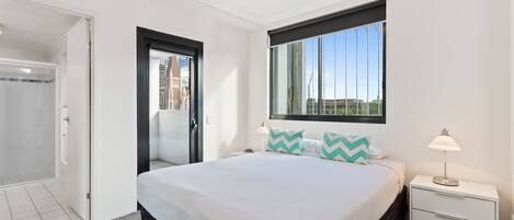 A hotel room with a bed, a nightstand, a window with a view of buildings, and a door leading to a shower area.