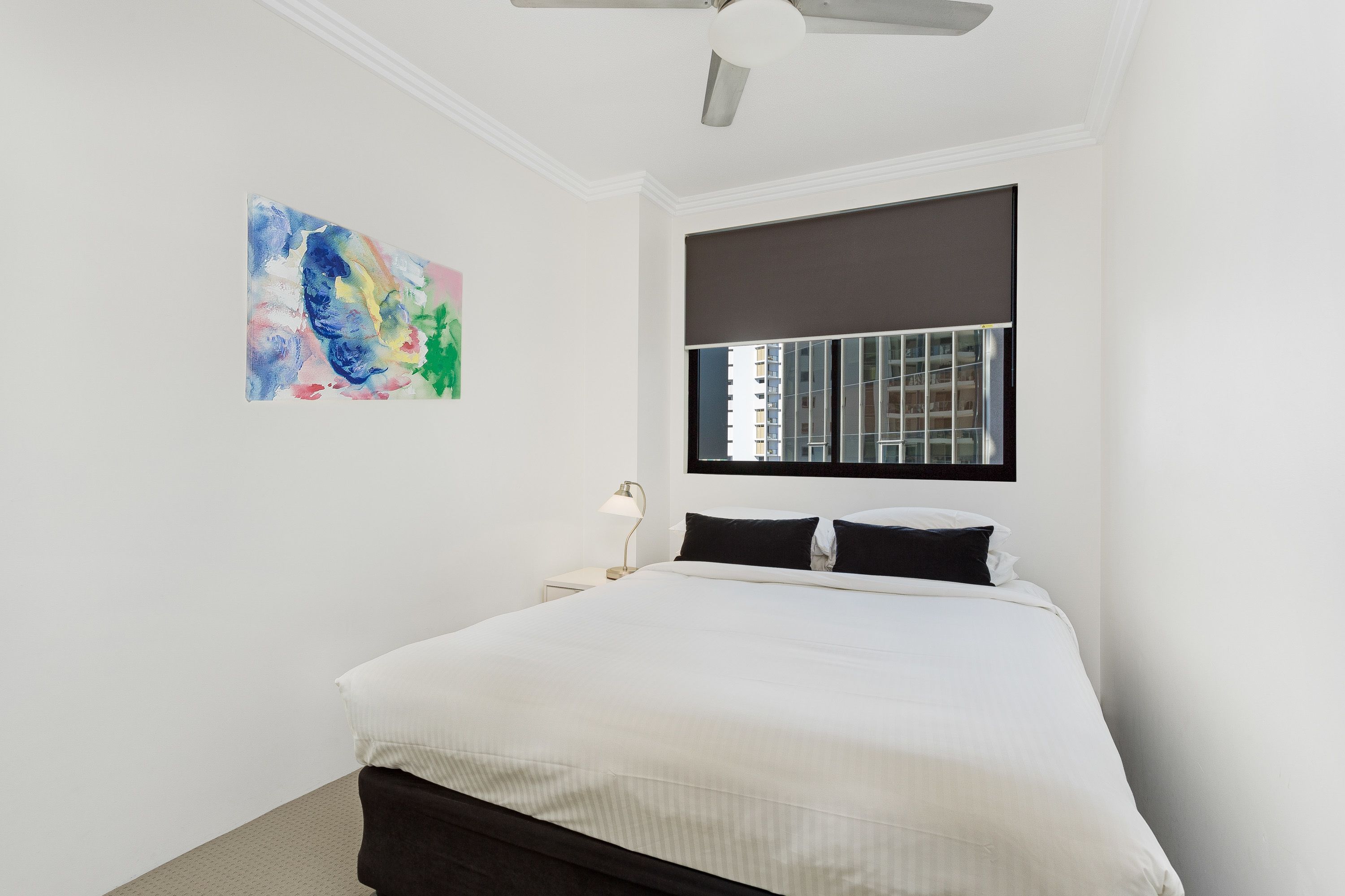 Une chambre à coucher avec un lit, une fenêtre avec des stores, un ventilateur de plafond et un tableau abstrait aux couleurs vives accroché au mur.