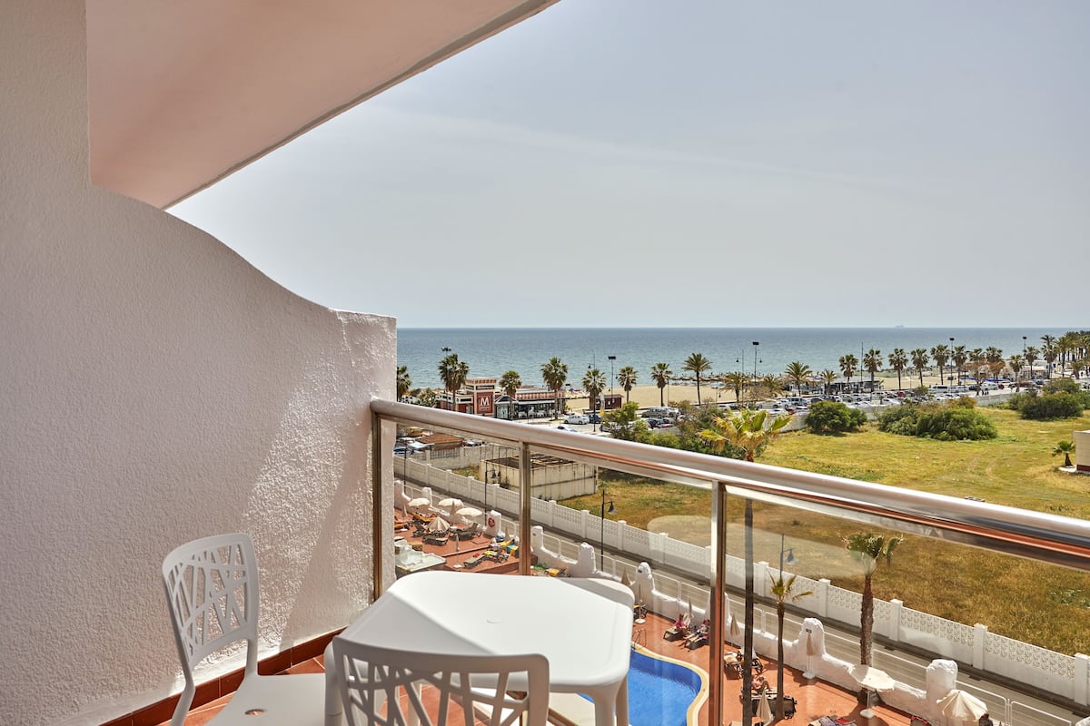 Un balcon avec vue sur la plage, une piscine et un espace repas aménagé avec du mobilier d’extérieur.