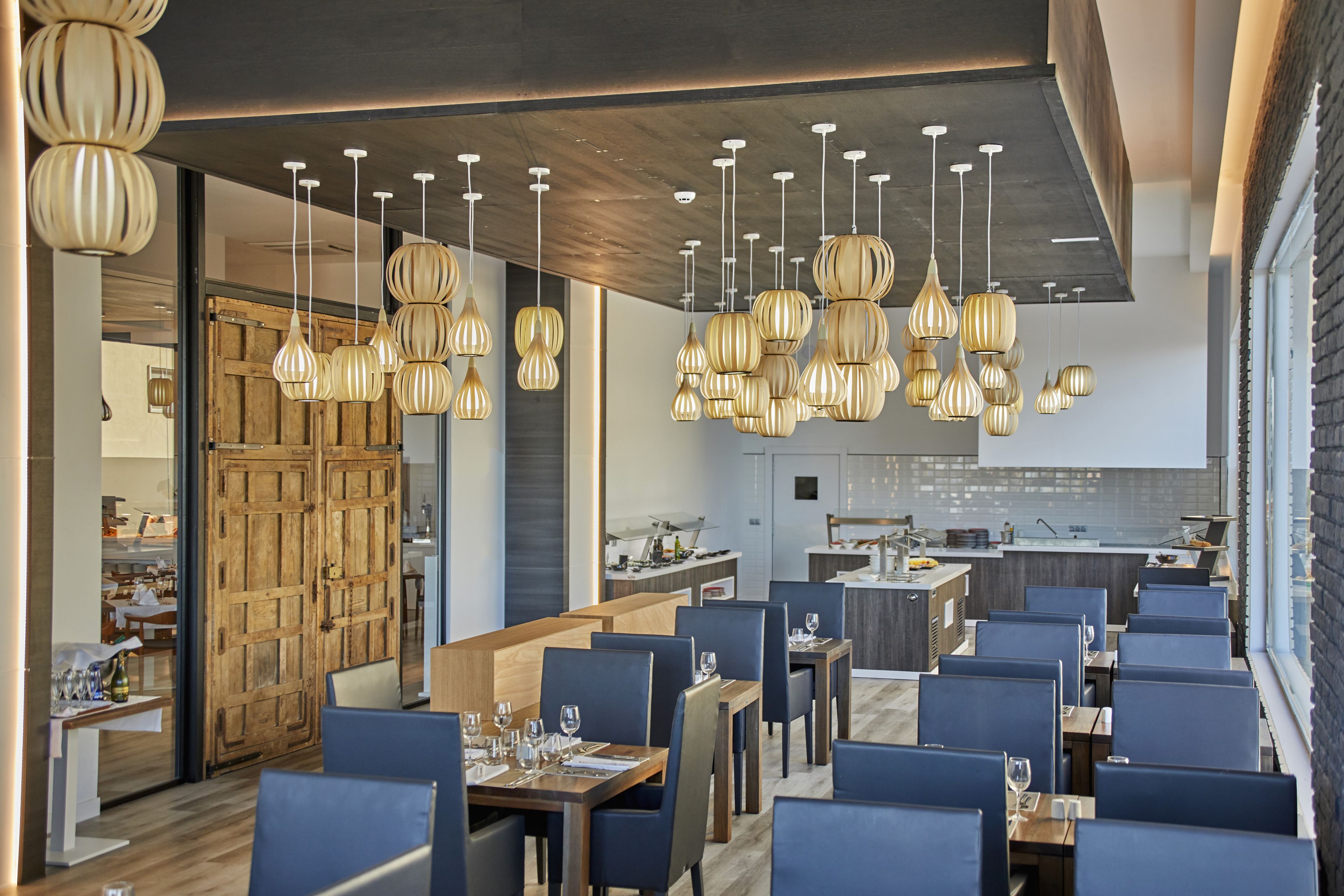 Een modern restaurantinterieur met houten tafels en blauwe stoelen, hangende lampen en een zichtbare keuken.