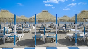 Een strand met rijen witte ligbedden en blauwe palen, waarvan sommige voorzien zijn van rieten parasols.