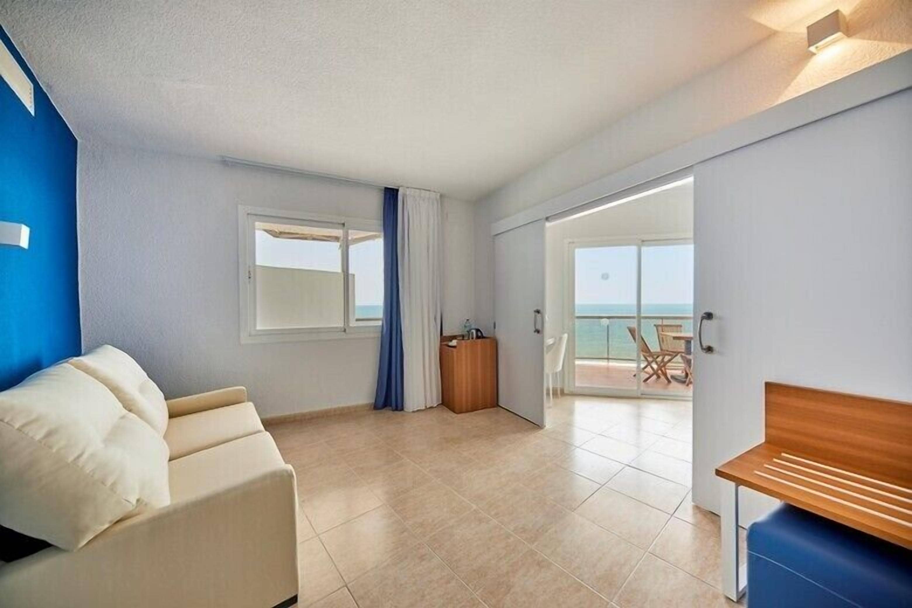 A modern hotel room with a sofa, a small table, a desk, and a balcony with a view.
