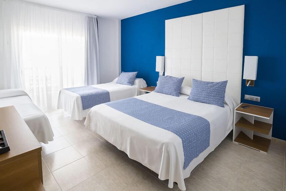 Une chambre d’hôtel avec deux lits, une grande fenêtre avec des rideaux, une table de chevet avec une lampe et une petite table en bois.