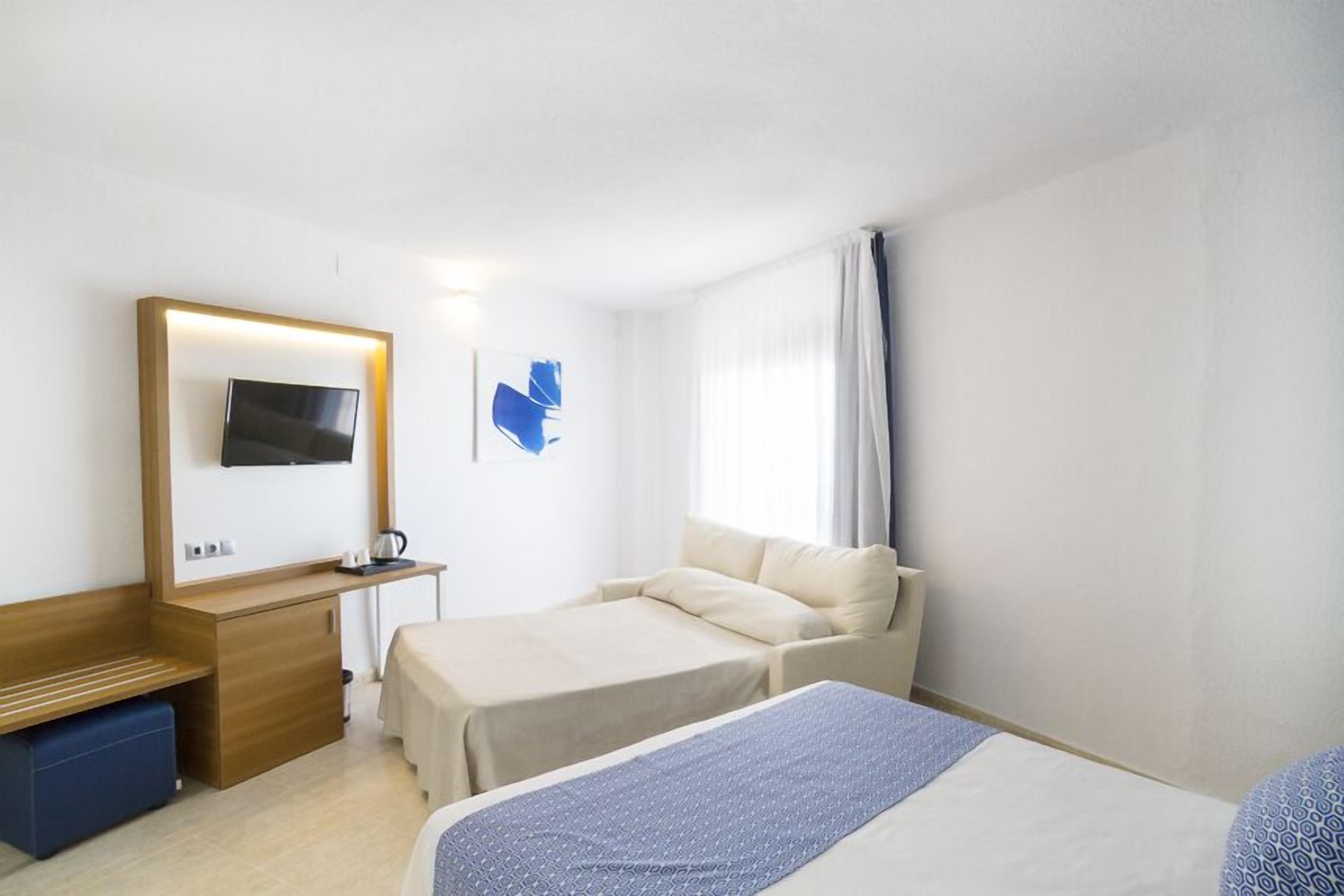 A hotel room with a bed, a desk with a TV, a small seating area, and a window with curtains.