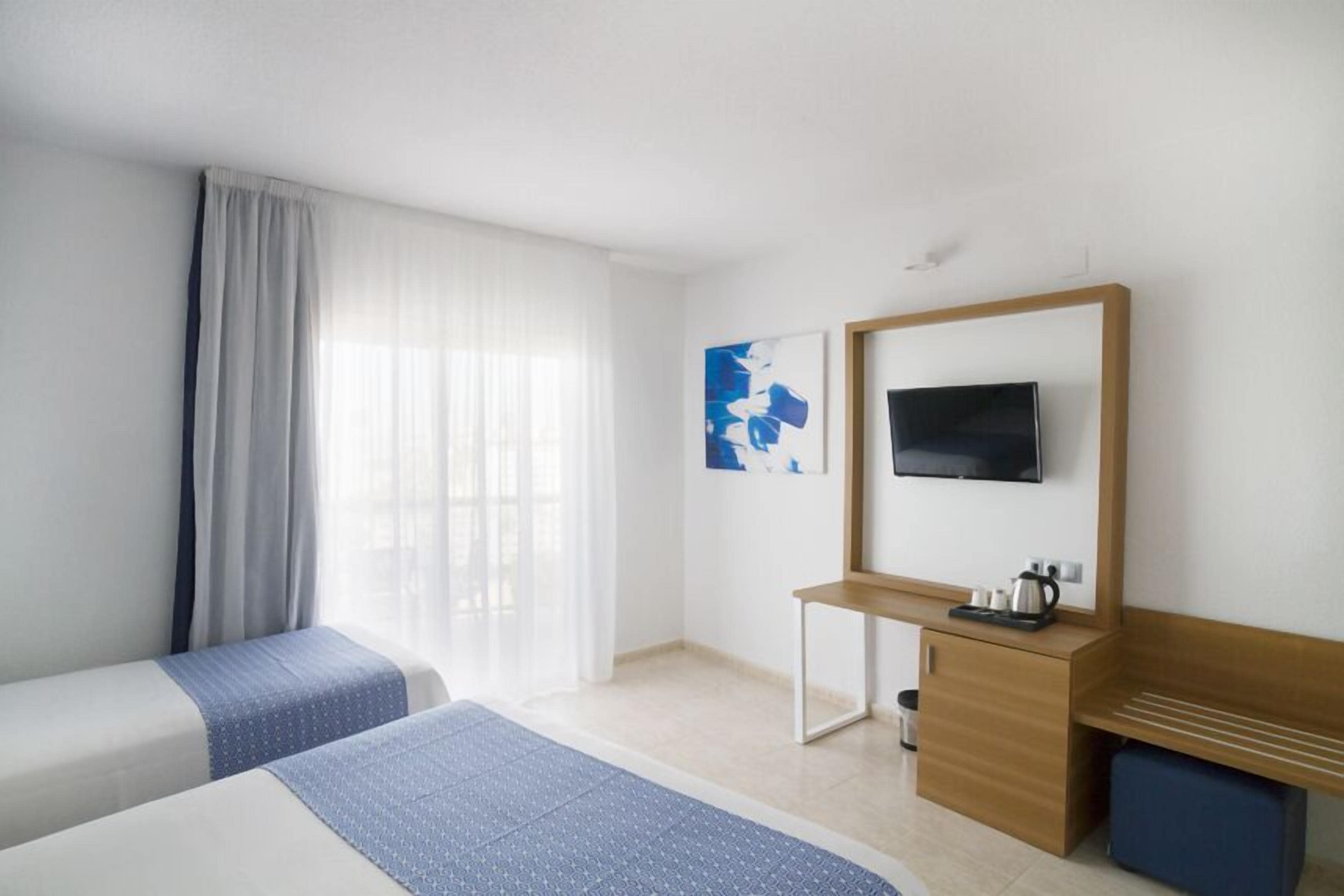 A hotel room with two beds, a desk, a television, and a window with curtains.