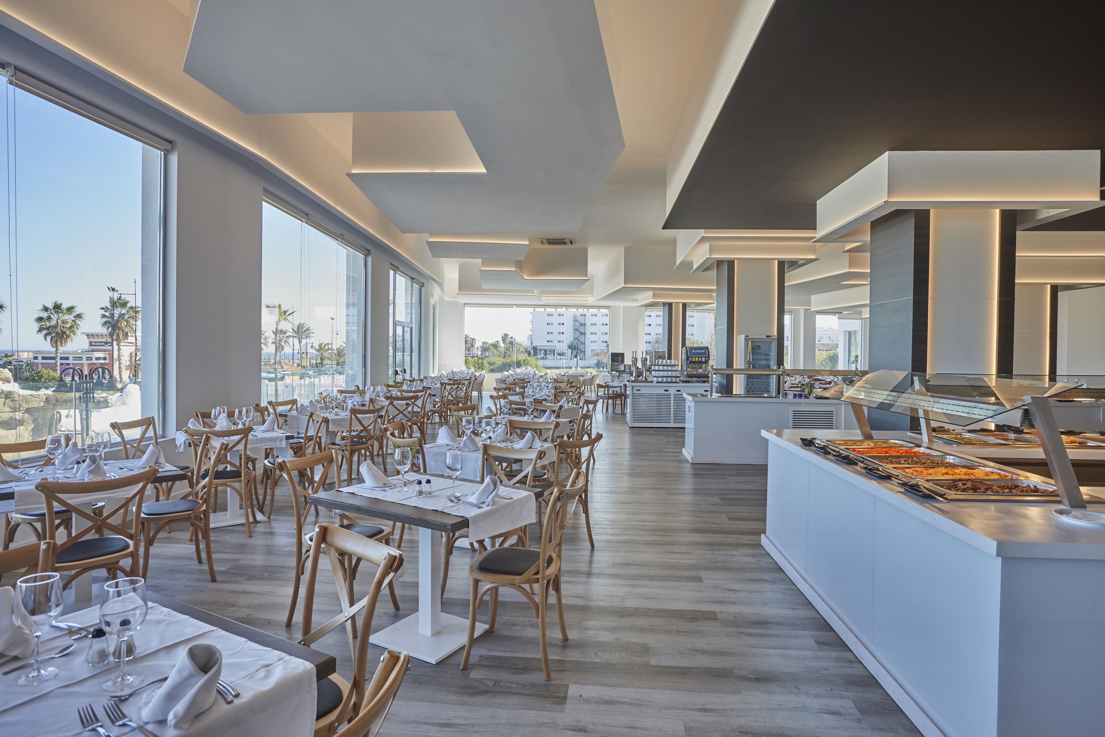 A spacious restaurant with high ceilings and large windows offering city views. Tables are neatly arranged with white tablecloths and wooden chairs.