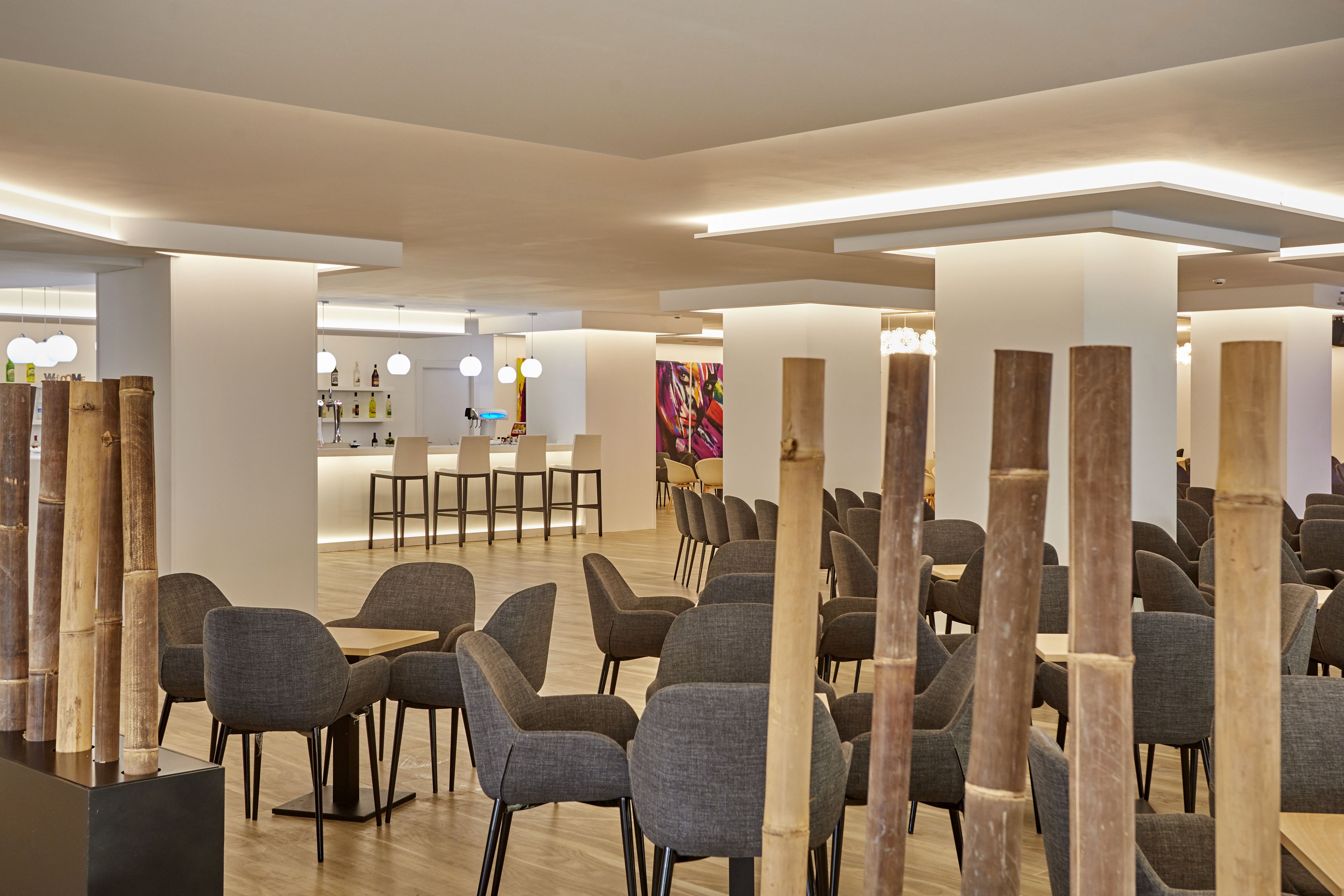 Un lobby moderno de hotel con filas de sillas y mesas grises, pilares de madera y un área de recepción al fondo.