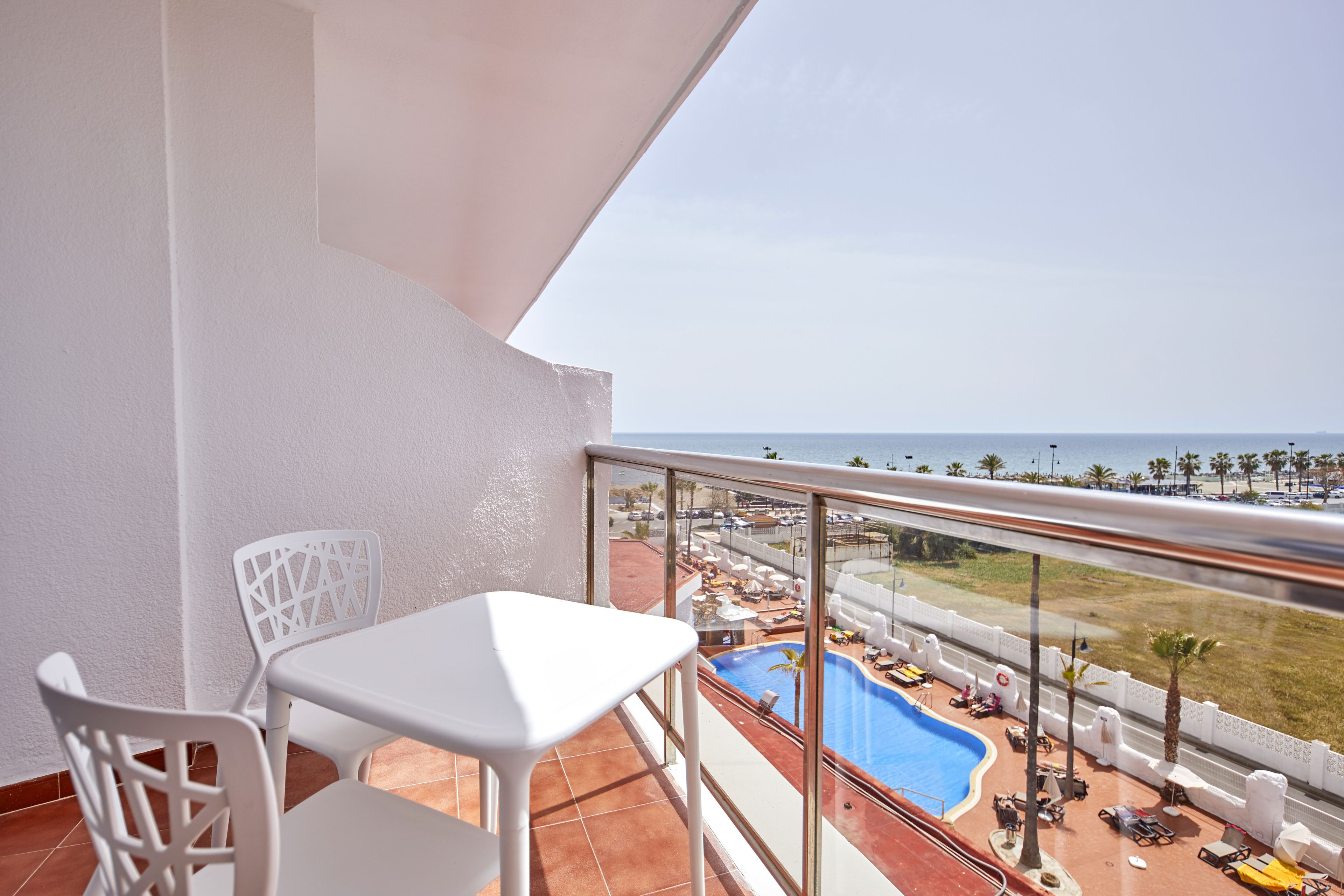 Un balcon avec une table et des chaises blanches, donnant sur un espace piscine équipé de chaises longues et de parasols, et offrant une vue sur l’océan.