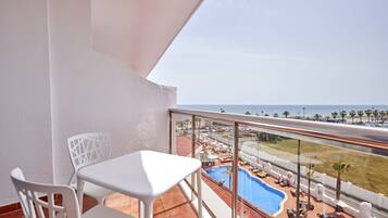 Un balcon avec une table et des chaises blanches, donnant sur un espace piscine équipé de chaises longues et de parasols, et offrant une vue sur l’océan.