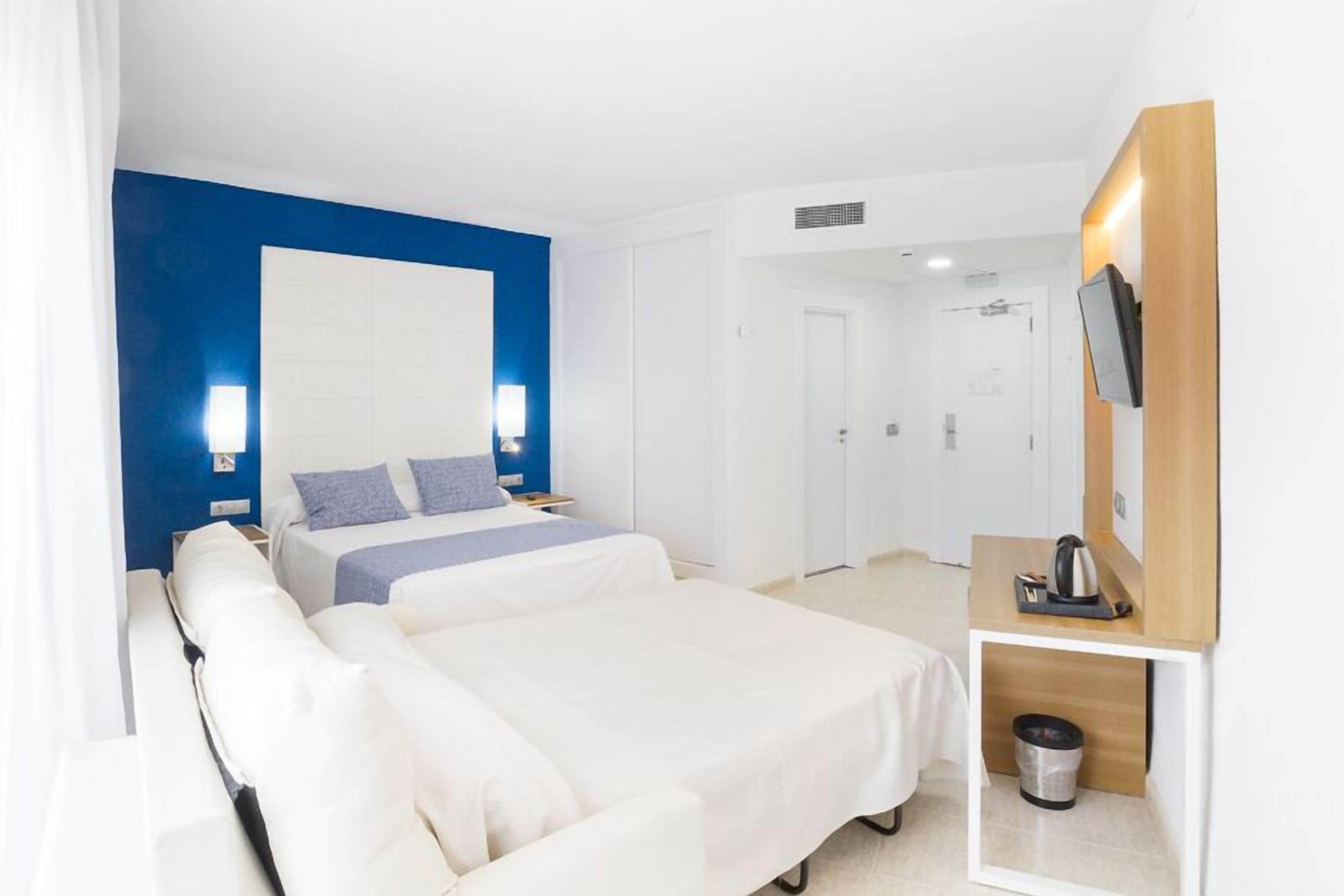A hotel room with two beds, a TV, a small table with a kettle, and a trash bin.