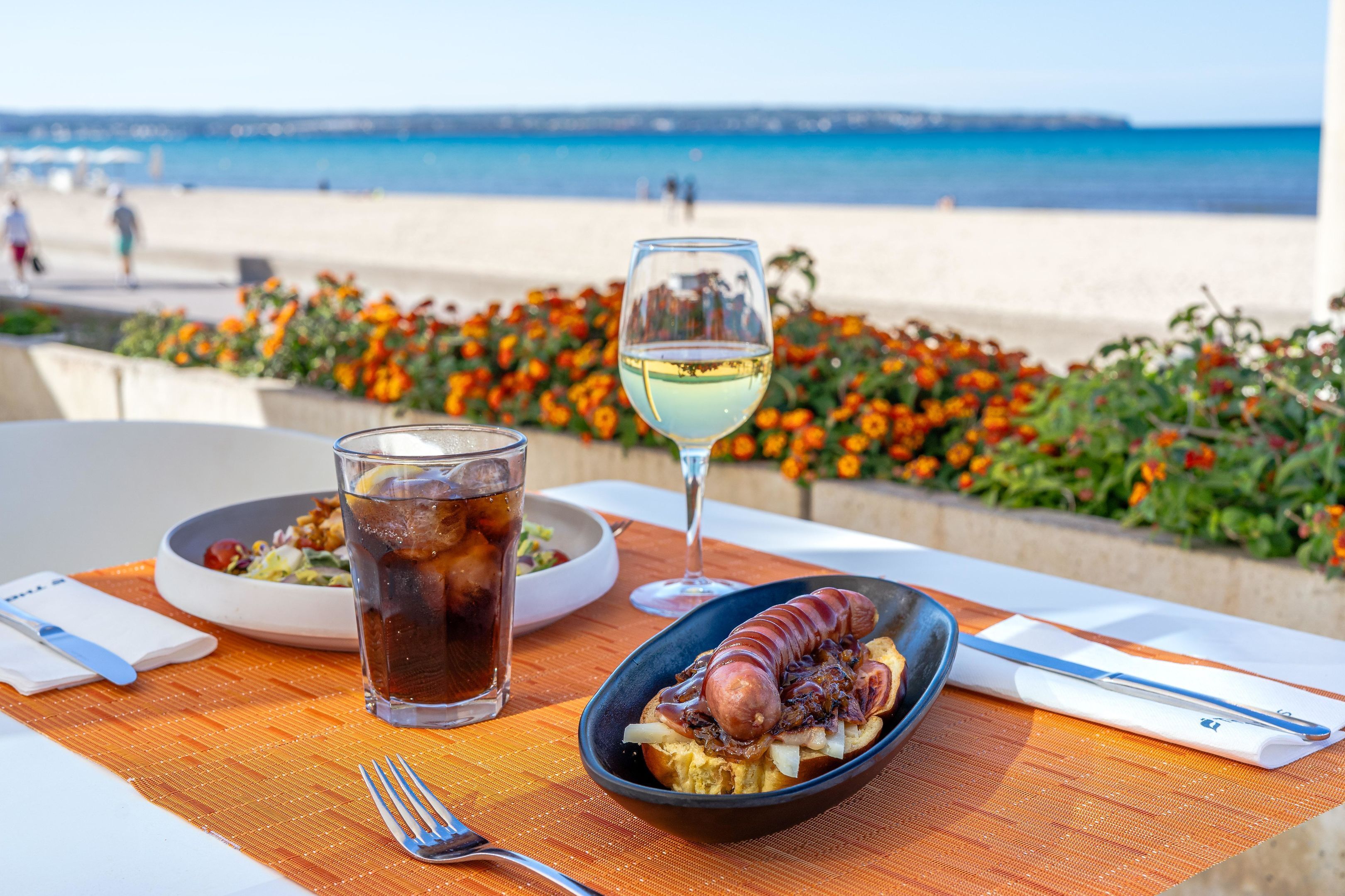 A meal with a glass of white wine and a drink on a table with a view of the beach.