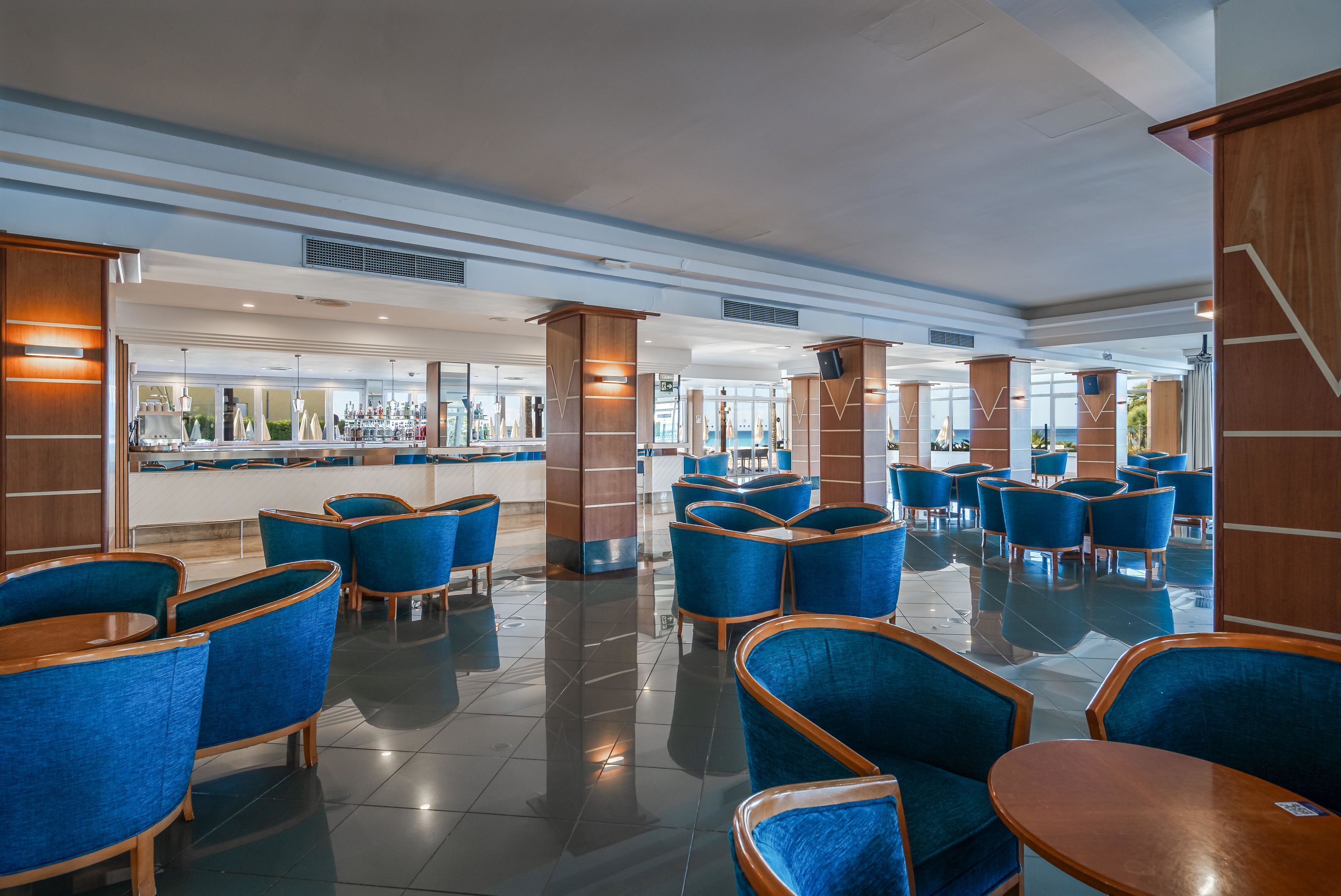 Un vestíbulo amplio con sillones tapizados en azul y mesas de madera, un área de bar al fondo y un diseño de techo moderno.