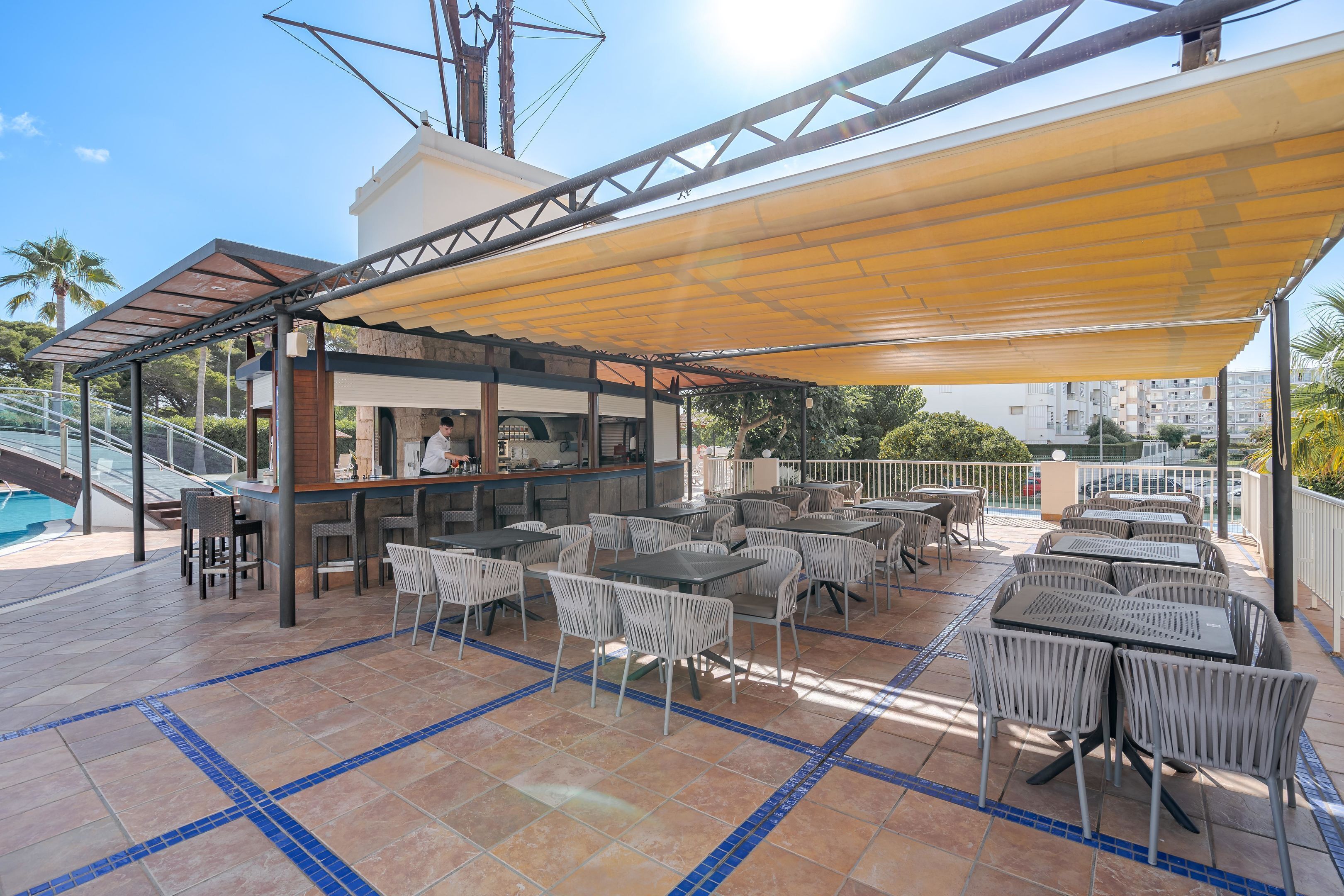 A covered outdoor seating area with tables and chairs, a bar, and a pool in the background.