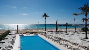 A pool area with lounge chairs and umbrellas, a clear view of the ocean, and palm trees.