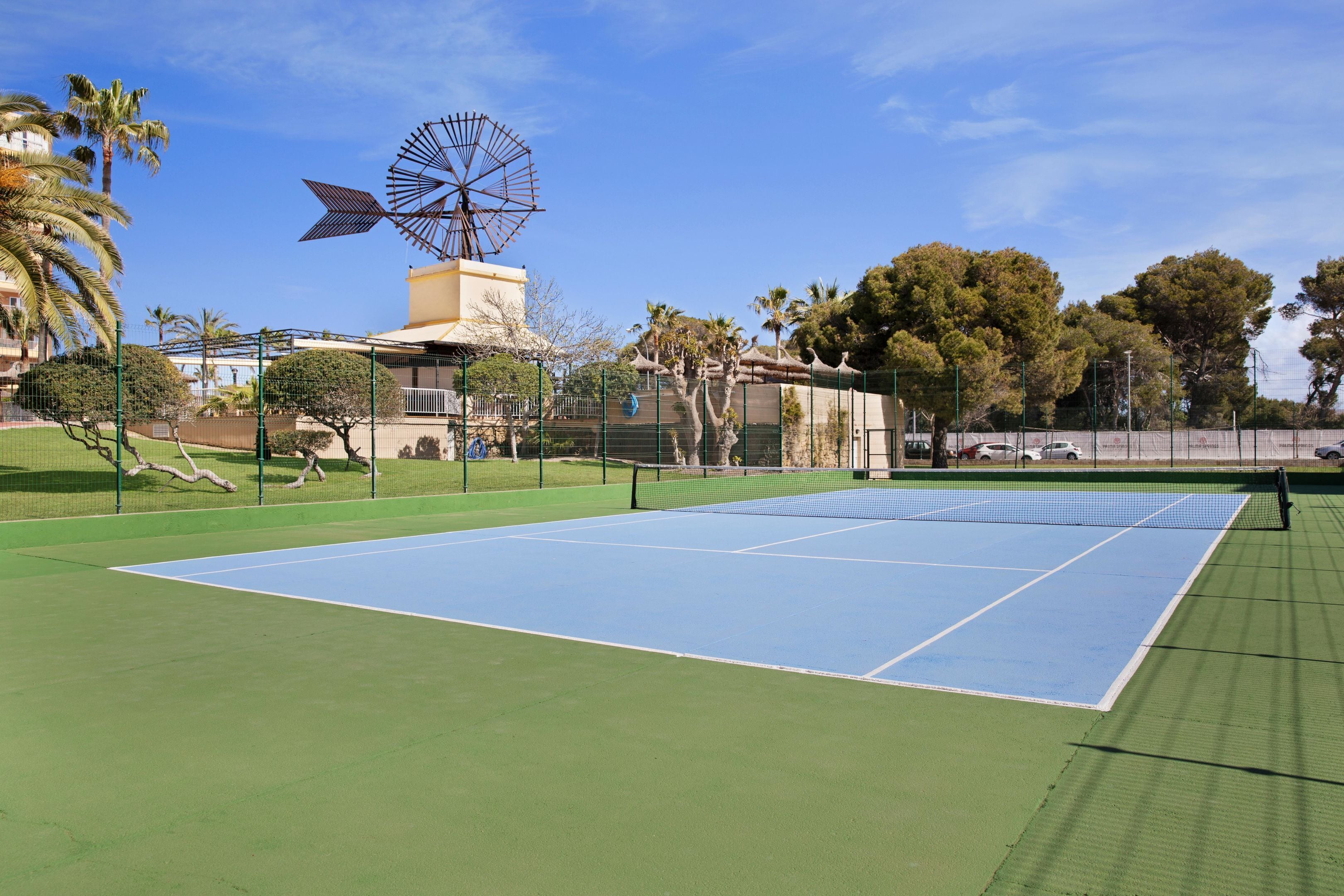 Cancha de tenis con un molino de viento al fondo, rodeada de palmeras y una valla.