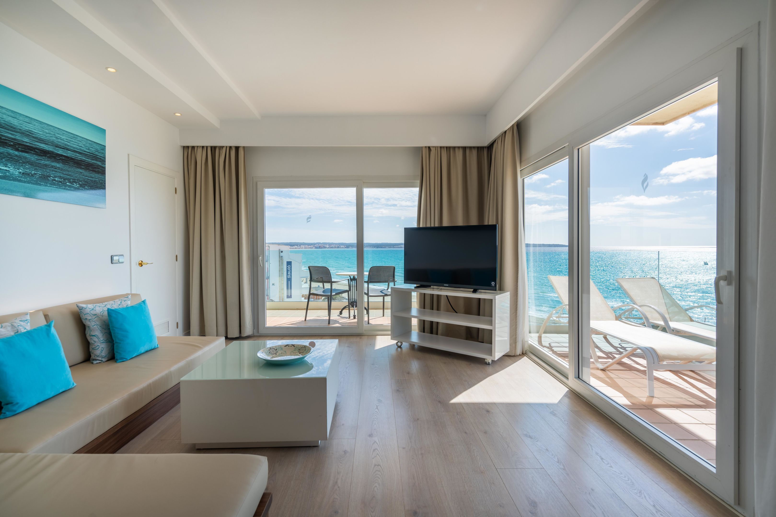 Un salon moderne doté d’un grand téléviseur, d’un canapé avec des coussins bleus, d’une table basse et d’un balcon offrant une vue sur la mer.