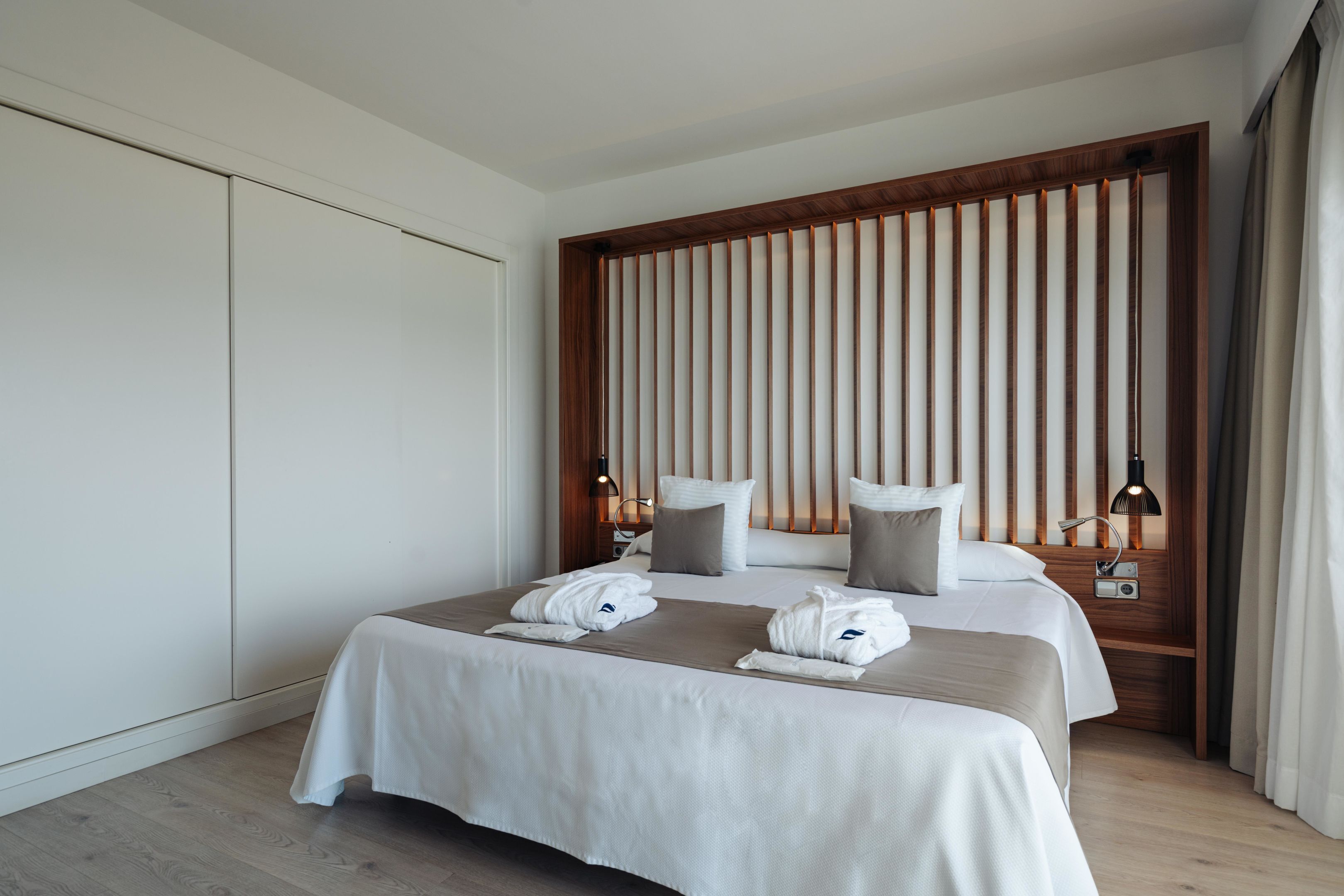 Une chambre d’hôtel moderne avec un grand lit, deux tables de chevet, une tête de lit en lattes de bois et une table de chevet avec une lampe.