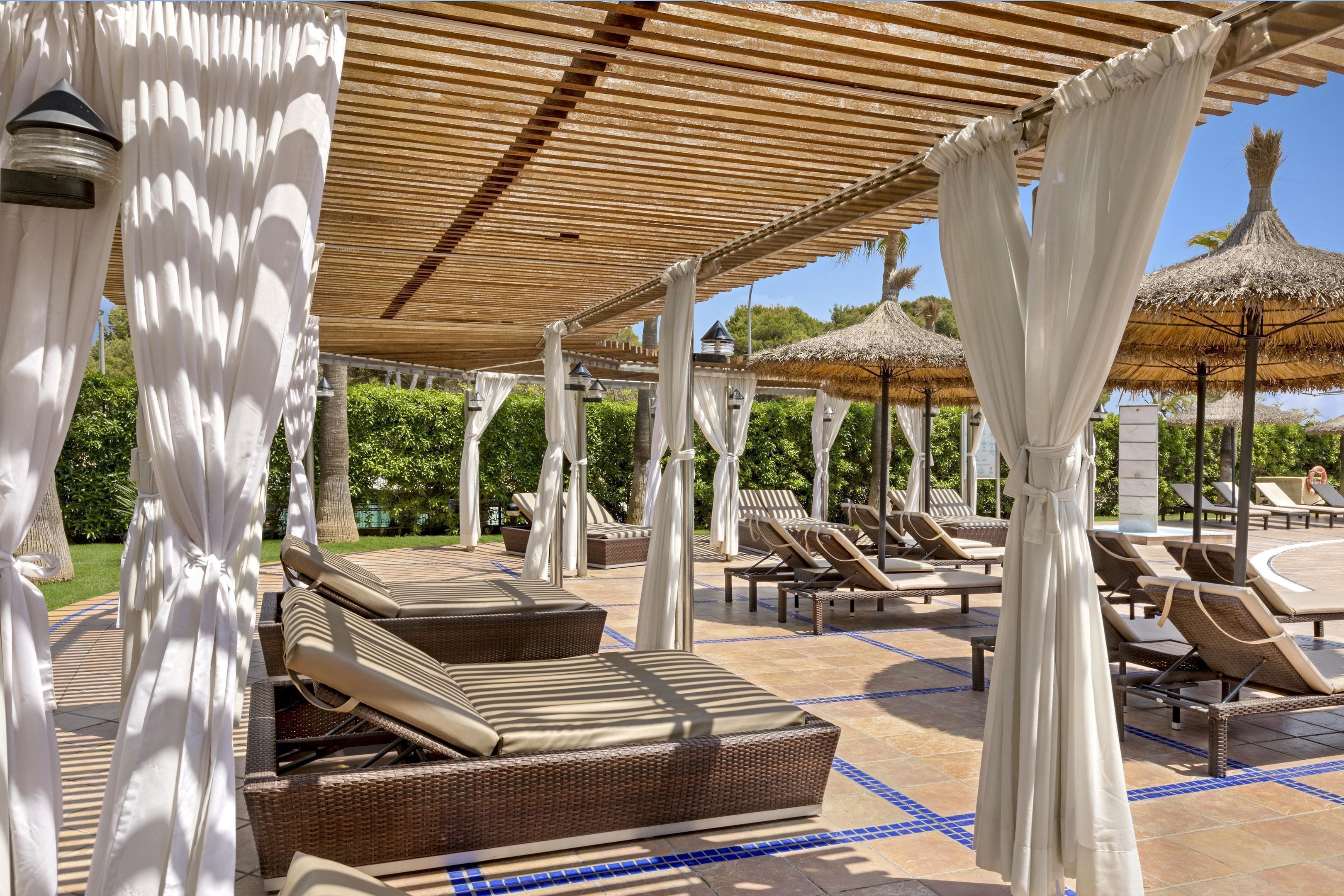 A covered outdoor lounge area with wicker furniture, white drapes, and thatched umbrellas.