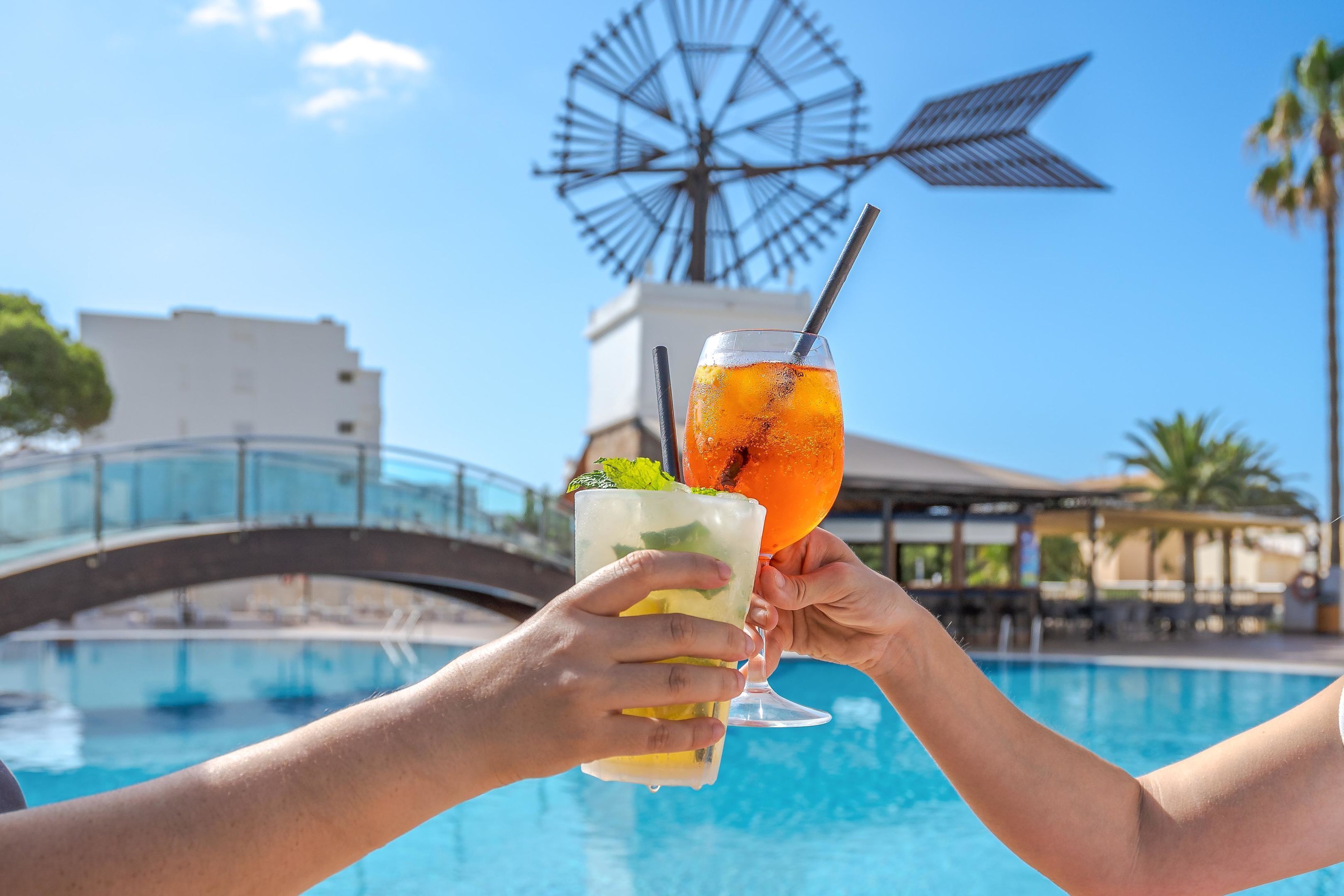 Two hands holding cocktail glasses with a windmill and a bridge in the background.