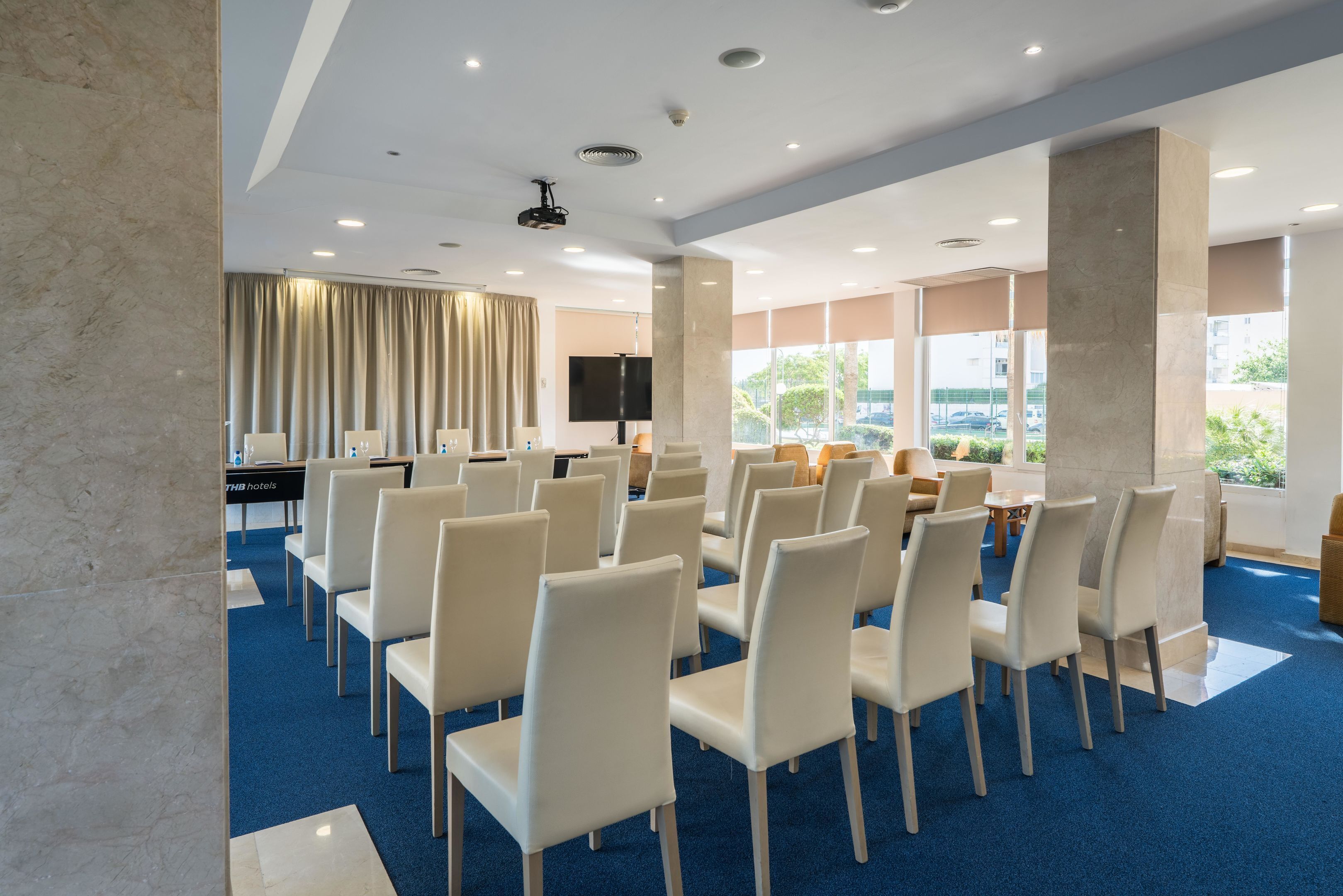 Un salón de conferencias con filas de sillas blancas, un escenario con un atril y un amplio ventanal con vistas al exterior.