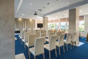 Uma sala de conferências com fileiras de cadeiras brancas, um palco com um púlpito e uma grande janela com vista para o exterior.