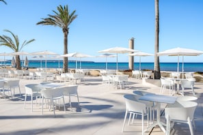 An outdoor café with white tables and chairs, palm trees, and umbrellas overlooking the sea.