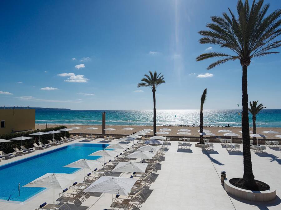 Un área junto a la piscina con sillones de descanso y sombrillas, con vista despejada a la playa y al océano, y palmeras.