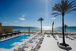 Un área junto a la piscina con sillones de descanso y sombrillas, con vista despejada a la playa y al océano, y palmeras.