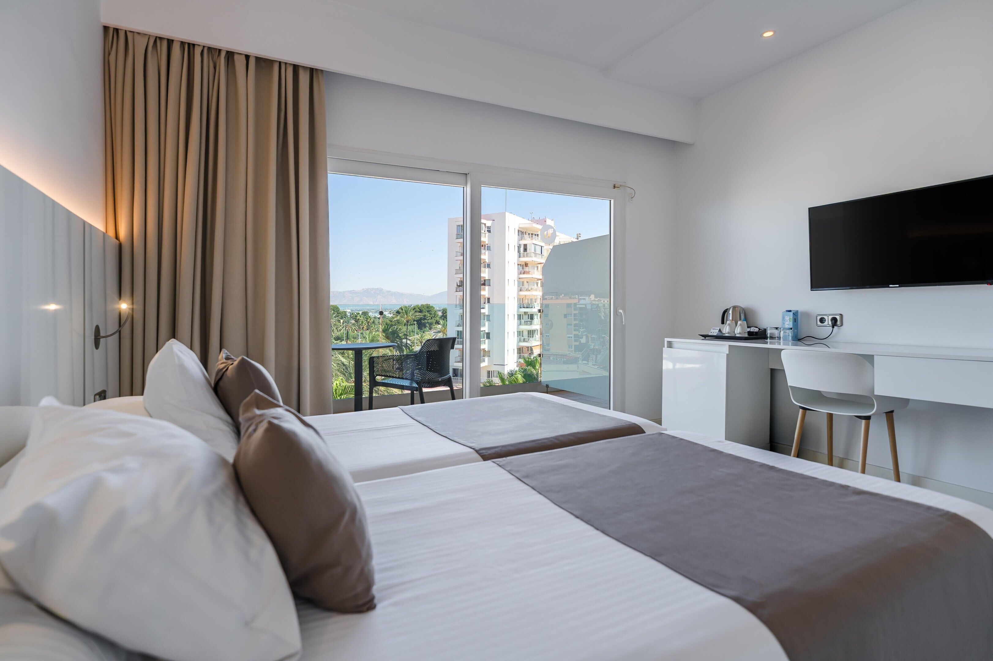 A hotel room with two beds, a desk, a chair, a TV, and a balcony with a view of buildings and greenery.